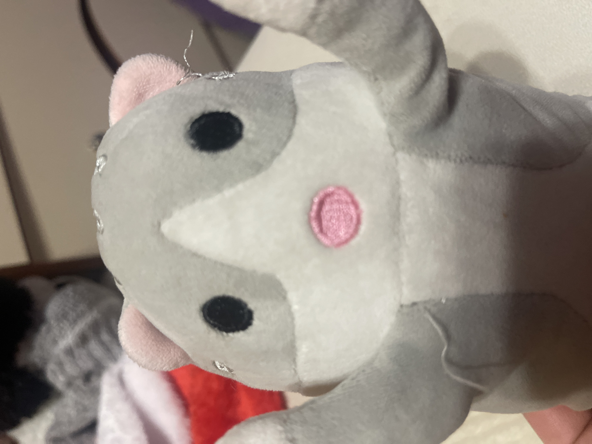 A close-up, slightly blurry photograph shows the front of a gray and white stuffed hamster toy with pink ears and a pink nose. The toy has black button eyes and its arms are raised. In the background, out of focus, are some gray, white, and red fabrics, and a brown surface. The scene is not located in the city of Minsk, Belarus.