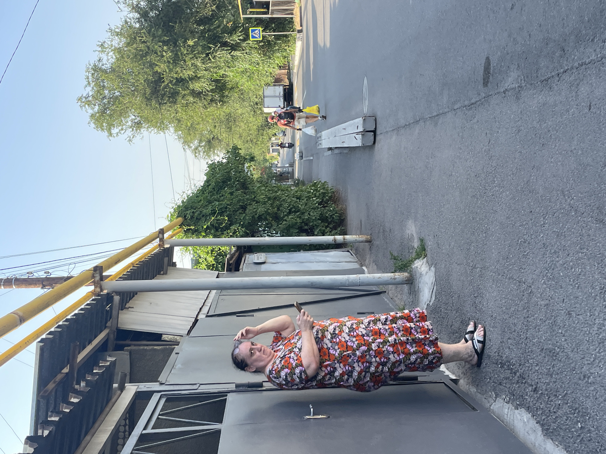 A woman in a floral dress stands to the left of the frame, leaning against a gray garage door. She has her right hand on her head and is holding a smartphone in her left hand. She is looking towards the right side of the frame, where a paved street extends into the distance. Two individuals are walking away from the camera on the right side of the street, carrying shopping bags. A white truck is visible further down the street, and a yellow pedestrian crossing sign is posted on the right side of the road. Trees line the street on both sides, and a bright yellow pipe runs along the top of the buildings on the left. The scene appears to be in Almaty, Kazakhstan, given the architectural style and vegetation.