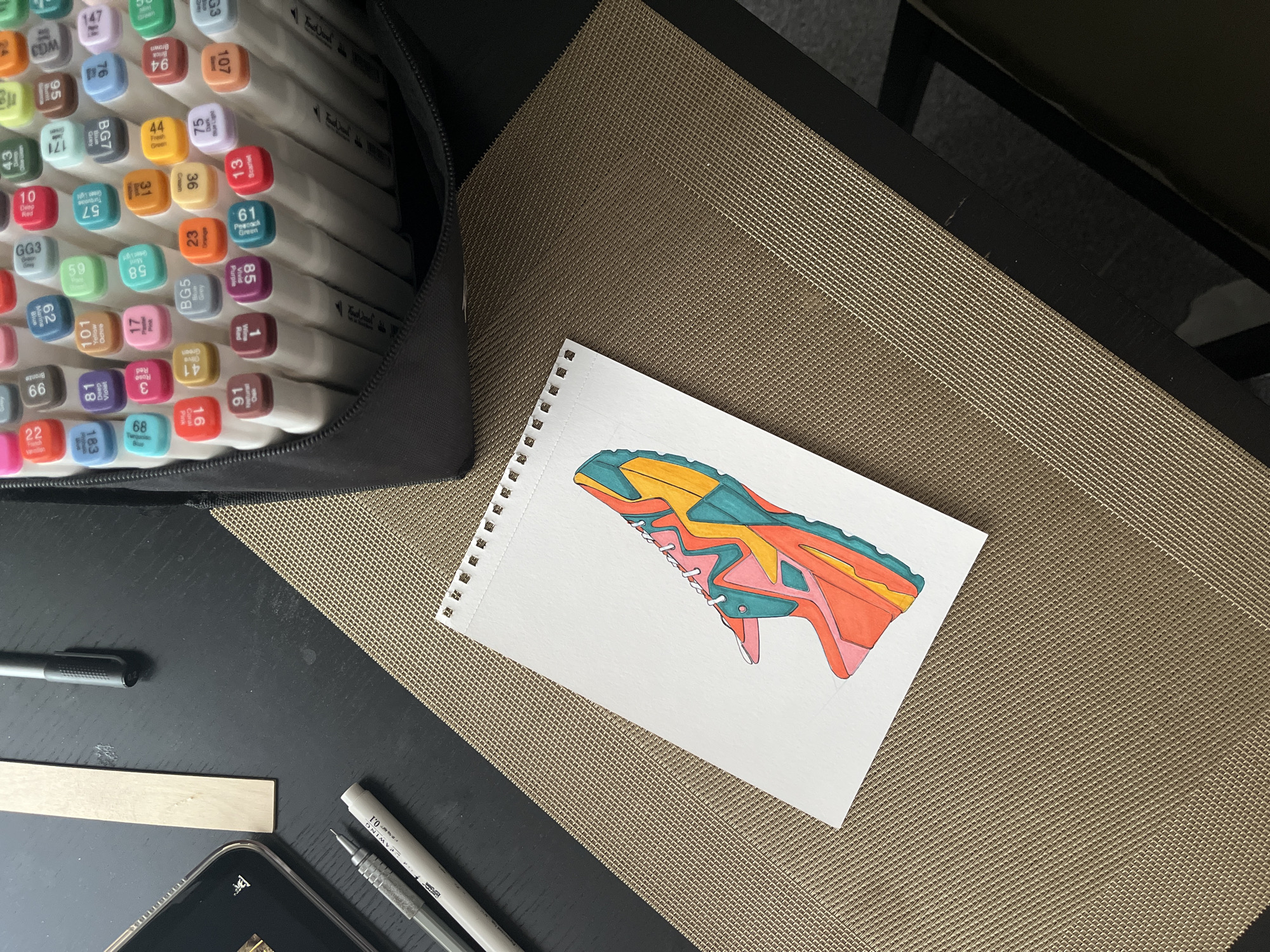 The image shows a workspace with art supplies and a drawing. A black zippered case filled with markers, identified by numerical codes like "63", "107", "94", and "147", is positioned at the top left. A white notebook with spiral binding contains a colorful drawing of a sneaker or shoe. A gray marker, a wooden ruler, a phone, and two pens are laid out near the notebook. The scene is set on a textured brown surface, likely a table or mat. No people or interactions are visible. The environment is simply an indoor space with art-related activities.
There is no indication about location in Krasnoyarsk, Russia.