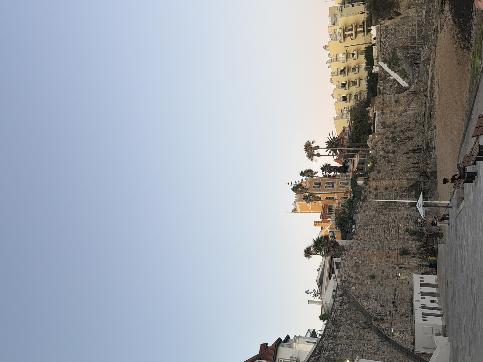 This media file captures a scene in Cascais, Portugal, during what appears to be late afternoon or early evening, with a clear sky. The image is oriented sideways.
The dominant subjects are buildings, a rocky cliff face, and a paved walkway. On the right side of the image, several people are visible. Two individuals are seated on a park bench, with one appearing to relax. A few other people are seen walking or standing near the cliff base, some in the vicinity of a white building with a tiled roof.
The scene depicts a coastal urban environment. Buildings of various architectural styles, including modern apartment blocks with balconies and older, more ornate structures, are nestled along the elevated terrain. The cliff face is composed of rough-hewn stone, with vegetation growing in crevices and along its edges. A sandy area, possibly a small beach or embankment, is present at the base of the cliff. A white flag with a circular emblem is mounted on a pole on the cliff.
Notable details include the stonework of the cliff, the design of the buildings, and the presence of palm trees suggesting a warm climate. The lighting indicates a transition from daylight to dusk. The overall atmosphere seems peaceful and relaxed, with people enjoying the outdoor space. No visible text is present in the image.