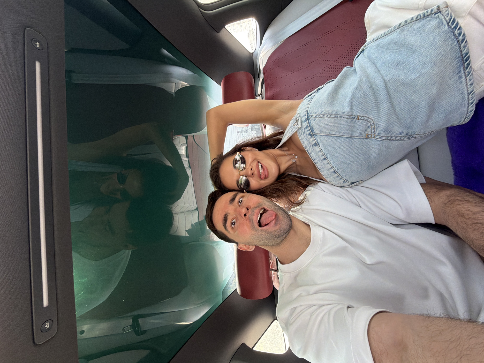 A selfie of a man and woman inside a vehicle in the Chaoyang District, China. The woman, wearing sunglasses and a white tank top with a light blue denim overall dress, sticks out her tongue. The man, wearing a white t-shirt, also sticks out his tongue. A clear glass roof reflects the scene. The interior features burgundy-colored seats. A control panel with a button featuring a person in a wheelchair is visible.