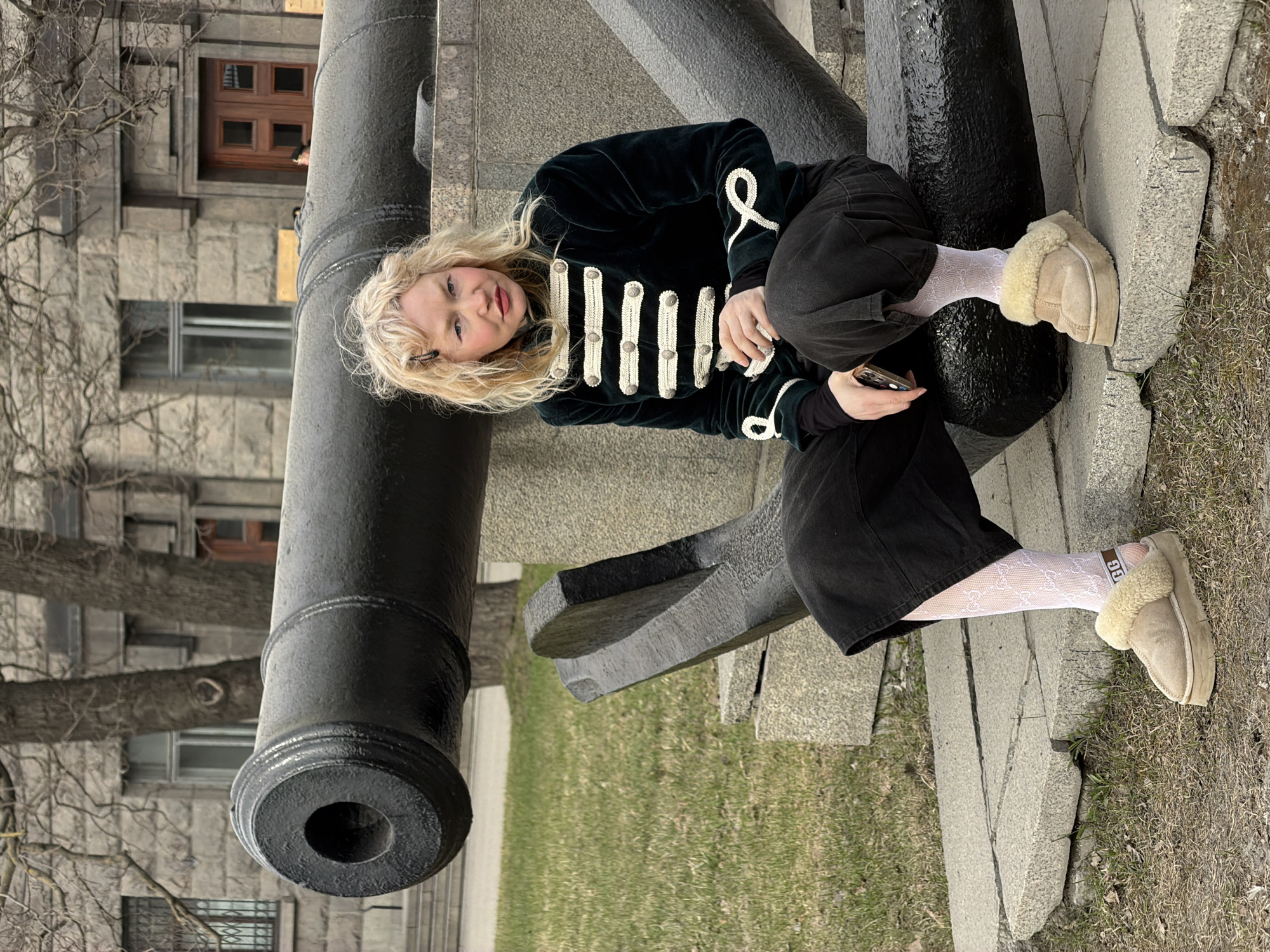 A woman is sitting on the barrel of a cannon. The cannon is located outside a building in Saint Petersburg, Russia. The woman has blond hair and is wearing a dark green velvet jacket with white braided details, black pants, white patterned tights, and beige fur-lined shoes. She is holding a cell phone. The cannon is black and sits on a concrete base surrounded by grass. A building with grey stone walls and windows is visible in the background, along with bare trees.