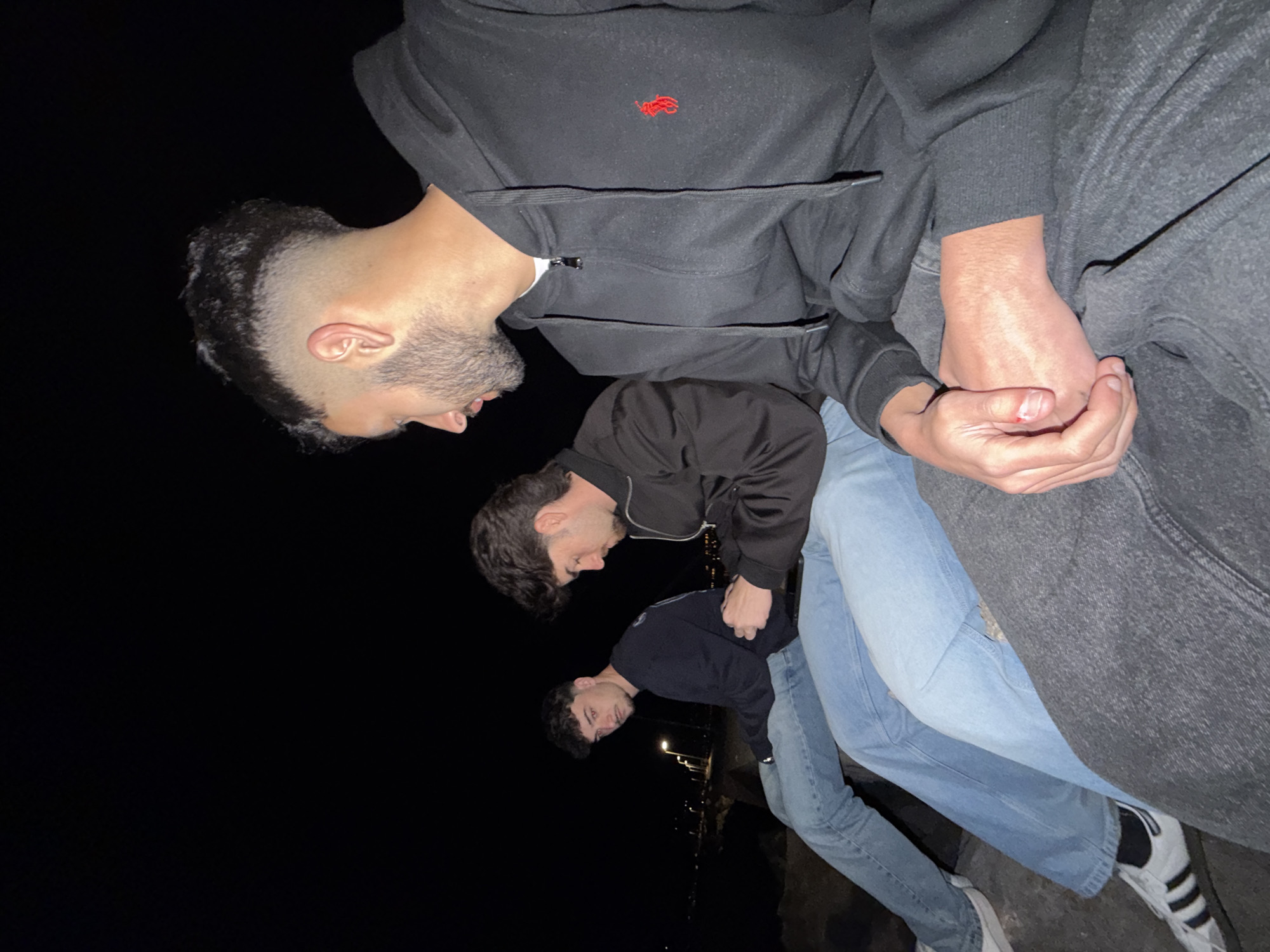 Three young men are posed closely together in an outdoor location in the city of Grad Rijeka, Croatia. The image is taken at night with the city lights visible in the background. The men are casually dressed in jeans and jackets/hoodies. The man at the top wears a black zip-up hoodie with a red logo, and gray sweatpants. His right hand is slightly injured. The other two men are wearing dark jackets and light-colored jeans. They seem to be standing on or near a stone ledge.