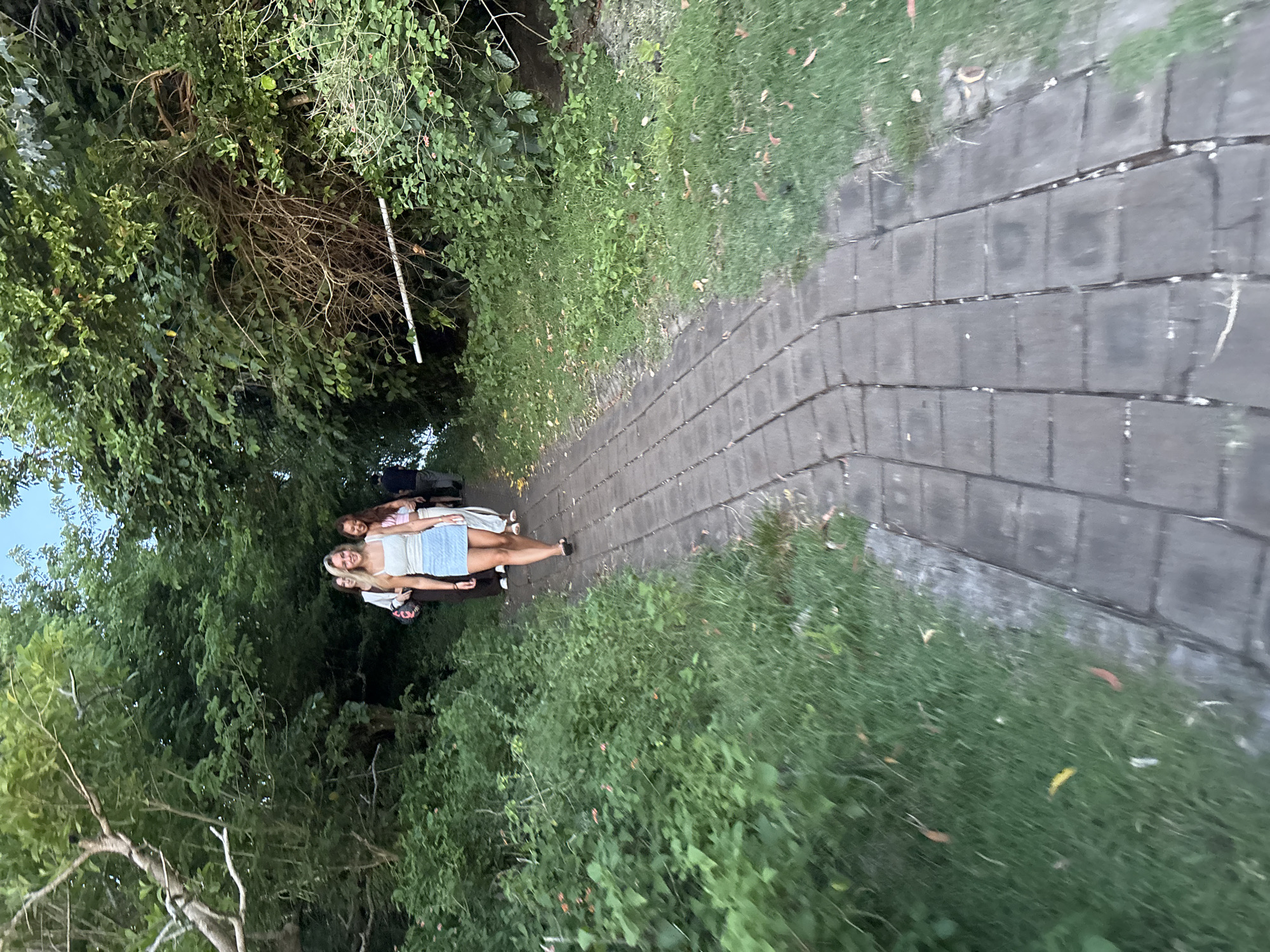 The image depicts a paved walkway lined with dense greenery, potentially in Uluwatu, Indonesia, given the tropical vegetation. Three individuals are walking along the path; the person in the foreground is fair-skinned with blonde hair, wearing a light-colored top and denim skirt. The individual behind them is fair-skinned with long red hair. The individuals appear to be on a leisurely stroll. The path is constructed of rectangular gray pavers. Vegetation, including trees and shrubs, borders both sides of the walkway, creating a tunnel-like effect.