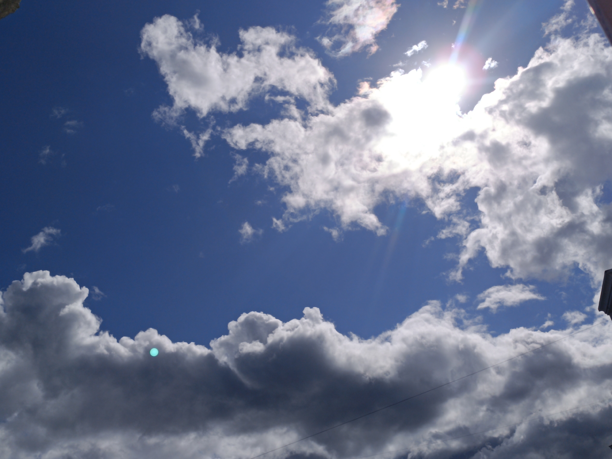 The image shows a bright blue sky filled with dynamic clouds, ranging from wispy white formations to dark, heavy storm clouds. The sun is high in the sky, creating a lens flare and illuminating the clouds from behind. One part of the sky has a distinct turquoise glow. There are some faint, thin lines that could be power lines or wires running horizontally across the frame. On the right edge of the image, the top of a dark structure, possibly a building, is visible. The scene appears to be an outdoor shot, likely captured during daylight hours.