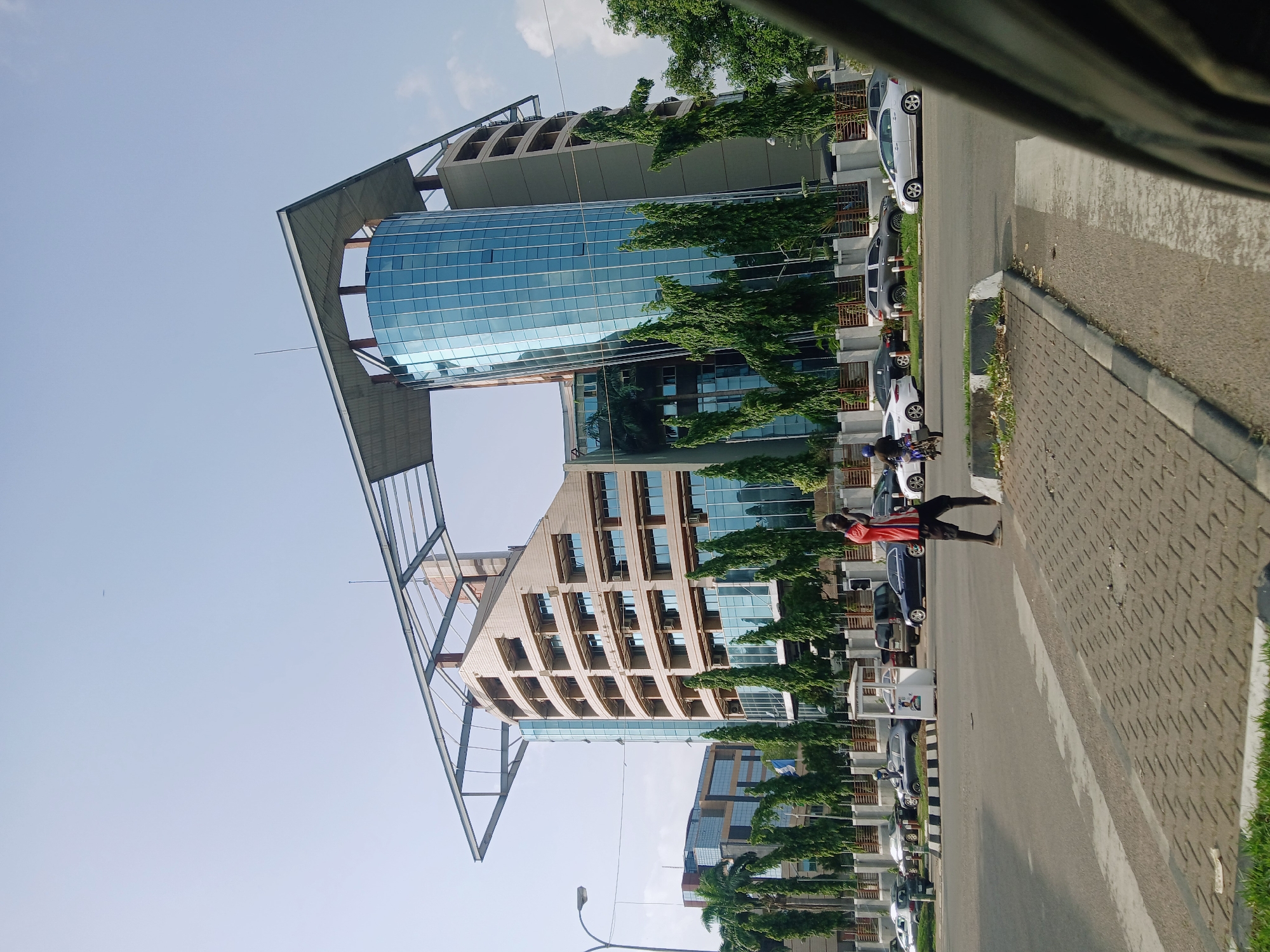 A person in a red top and black shorts is walking on a paved sidewalk next to a street lined with parked cars and trees. A large, modern building with a glass facade and a distinctive metal framework is visible in the background. The building's design incorporates both curved and angular elements. A few parked cars are seen along the street. The scene appears to be located in a business or commercial district in Abuja, Nigeria.