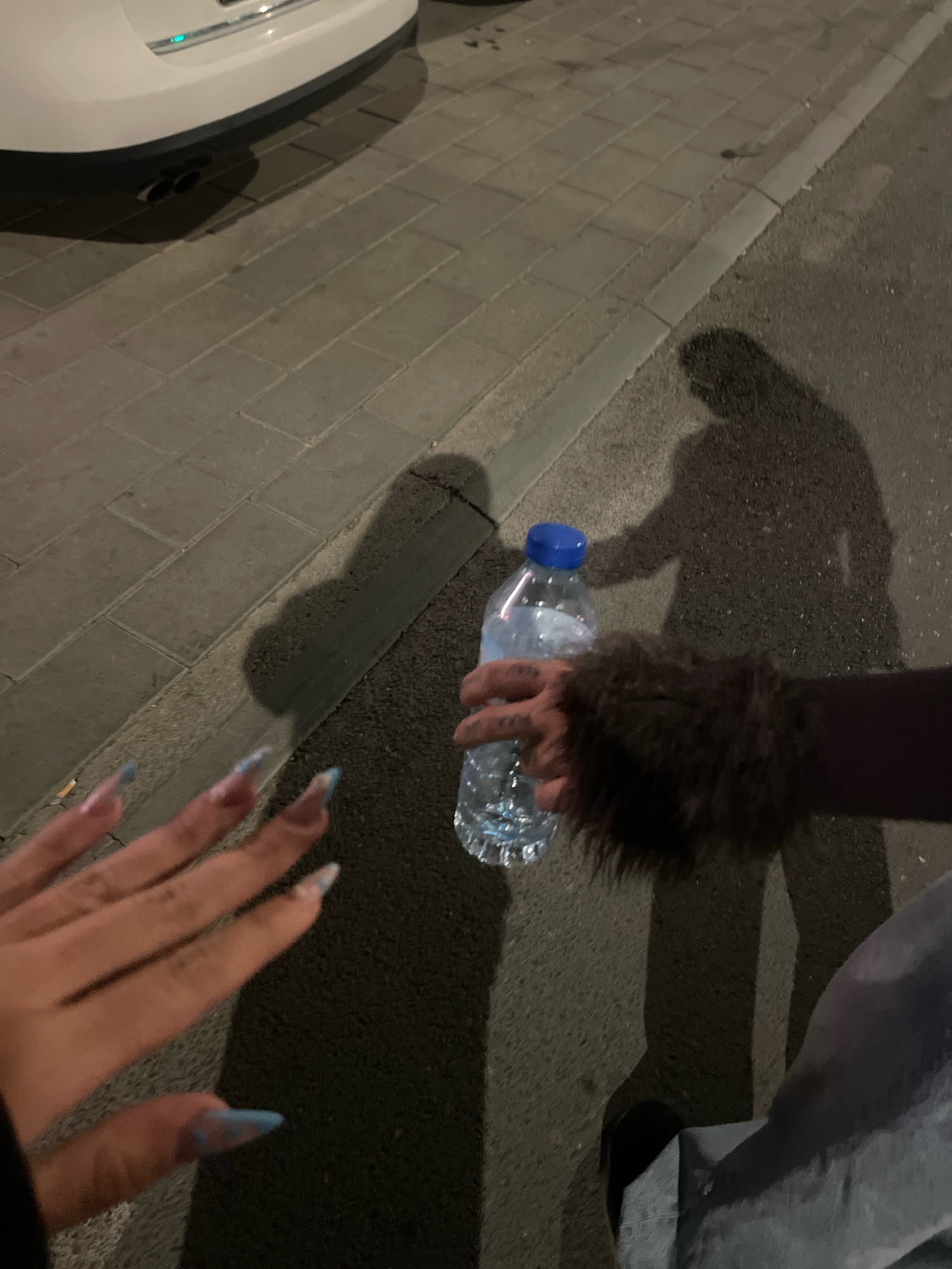 The image depicts a ground-level view of a paved sidewalk and asphalt street, illuminated by artificial light. A white car is partially visible in the upper left corner, with its exhaust pipes showing. A shadow of a person holding a water bottle is cast on the street. Another shadow, seemingly of a second person, is also visible. In the foreground, a person's hand with long, light blue-tipped fingernails is held up, positioned over the sidewalk. A dark, furry accessory is visible on the arm of the person holding the water bottle. The scene appears to be outdoors, in a city environment. The mention of Prizren, Kosovo, suggests this is a potential location for the image.