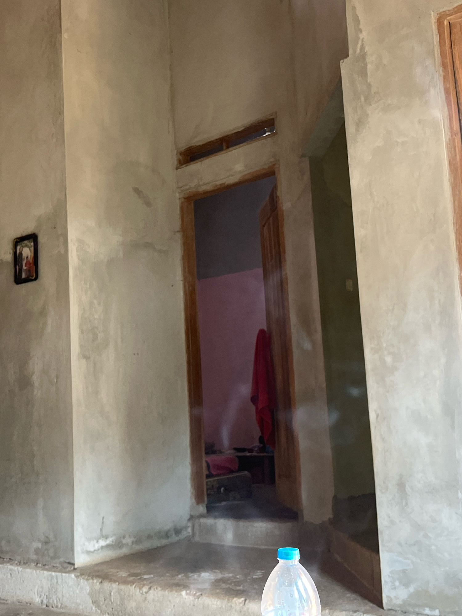 The image depicts an interior scene with concrete walls and a wooden doorway leading into a room with pink walls and a red cloth hanging. In the foreground, a plastic water bottle with a blue cap is visible. A small framed picture hangs on the left wall. The scene appears to be an unfinished or sparsely furnished building. The location is identified as Sinar Mulyo, Indonesia.