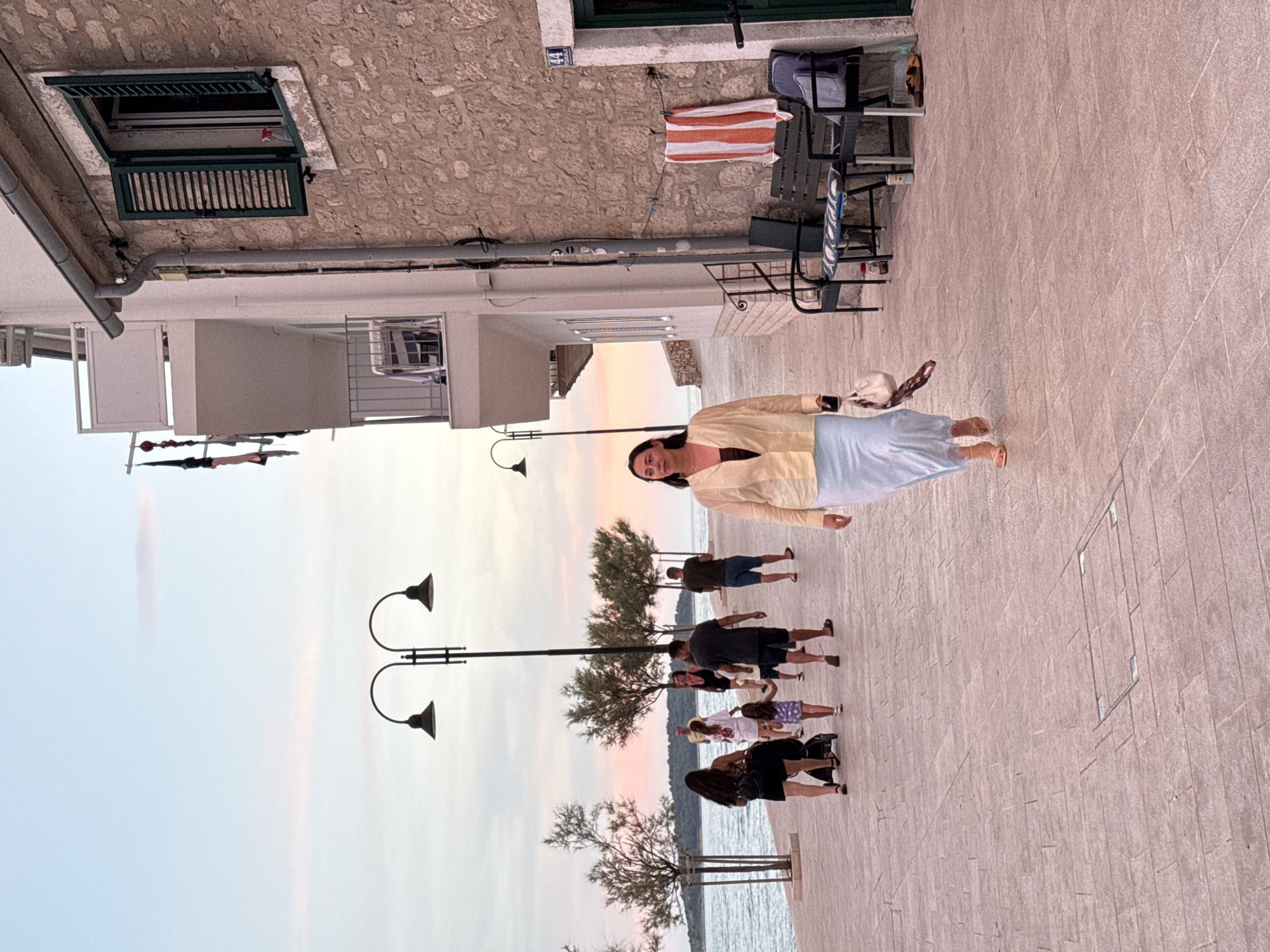 The image shows a woman walking on a paved waterfront promenade in Primošten, Croatia. The woman is wearing a light yellow blouse and a light blue skirt, and she is carrying a beige handbag. She is walking towards the viewer. In the background, several other people are walking along the promenade, some with children and a stroller. There are trees lining the promenade, and beyond them, the sea. The sky is a soft pink and orange, suggesting it is sunset. To the right of the woman is a building with a stone facade and a green shuttered window. There are benches and a towel hanging on the wall of the building. The street number "44" is visible above a doorway.