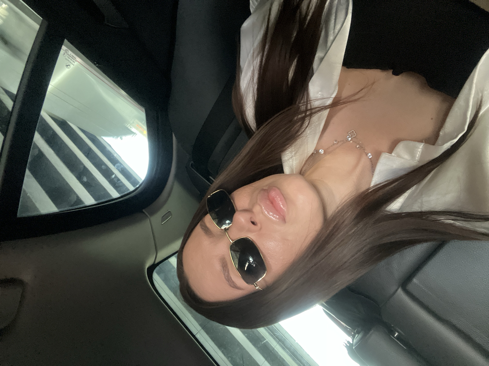 A woman wearing sunglasses and a white blouse is in the back seat of a car. The car's window shows an interior shot of a parking garage. The location cannot be determined from the image.