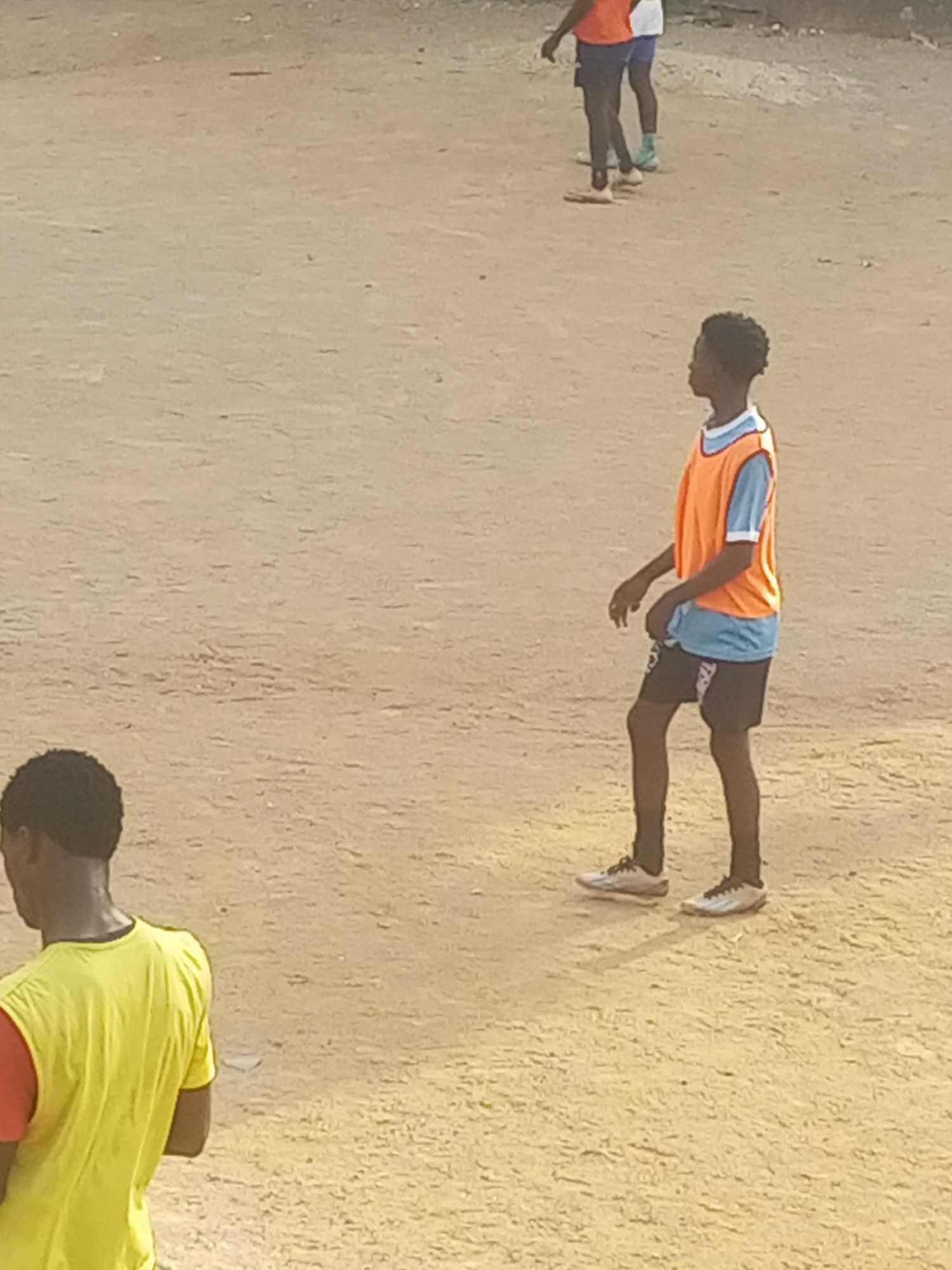 The image depicts an outdoor scene on a dirt or sandy surface, likely a field or court. In the foreground, a person wearing a yellow t-shirt and a red undershirt is partially visible from the back, facing away from the camera. To the right, a young person wearing an orange bib over a blue t-shirt and black shorts is standing and looking to the left. Further in the background, two other individuals are visible, one wearing a blue and white outfit and the other in a similar orange bib and blue shorts. The environment appears to be an open, unpaved area with a light brown, dusty surface. There are no discernible landmarks or specific city features to identify the location.