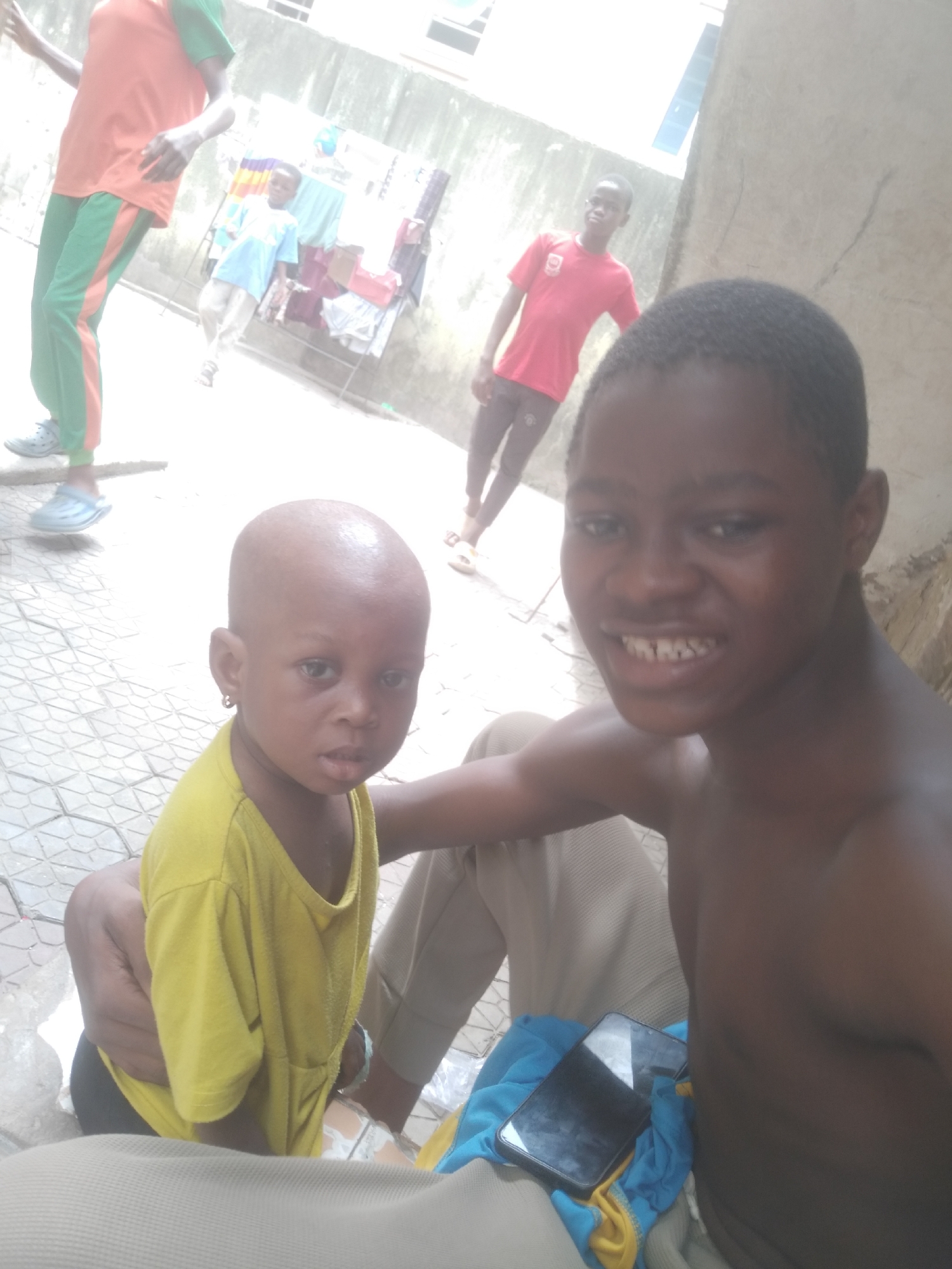 The image captures a casual outdoor scene featuring several dark-skinned individuals in what appears to be a residential or communal area in Shangisha, Nigeria, during bright daylight.
In the foreground, a shirtless young male, likely a teenager, is on the right, smiling broadly towards the camera. His arm is around a bald toddler in a yellow top and dark bottoms, who is on the left and looks directly at the camera with a neutral, inquisitive expression. The two are positioned on a tiled ground with a geometric pattern.
In the midground, partially visible on the left, is a person wearing an orange t-shirt with green sleeves and matching green-striped pants, along with light blue Crocs-style shoes. Further back, a small child in a light blue shirt stands near a metal clothes drying rack laden with various garments. On the right side of the background, another young male in a red t-shirt and dark pants is smiling.
The background consists of light-colored concrete walls, suggesting an enclosed outdoor space, possibly a courtyard or an alleyway between buildings. A black smartphone rests on a blue fabric or towel next to the shirtless male in the foreground. The bright, somewhat washed-out lighting indicates it is daytime, likely under direct sun. The expressions on some faces suggest a friendly and relaxed atmosphere. No visible text is discernible in the image.