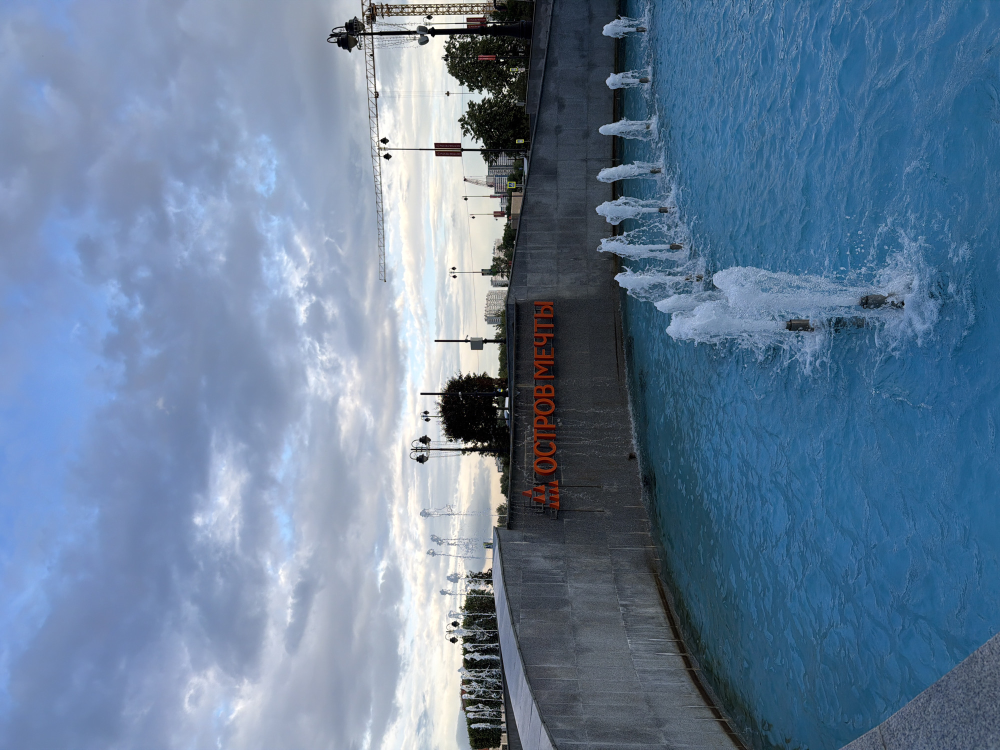 The image shows a public plaza with a fountain in the foreground. The fountain has bright blue water and multiple jets of water are spraying upwards. Behind the fountain is a grey, curved concrete wall with orange lettering that reads "ОСТРОВ МЕЧТЫ". Beyond the wall, there are trees, street lamps, and buildings in the distance. A construction crane is visible in the upper right corner of the image, silhouetted against a cloudy sky. The overall scene depicts an outdoor urban environment.