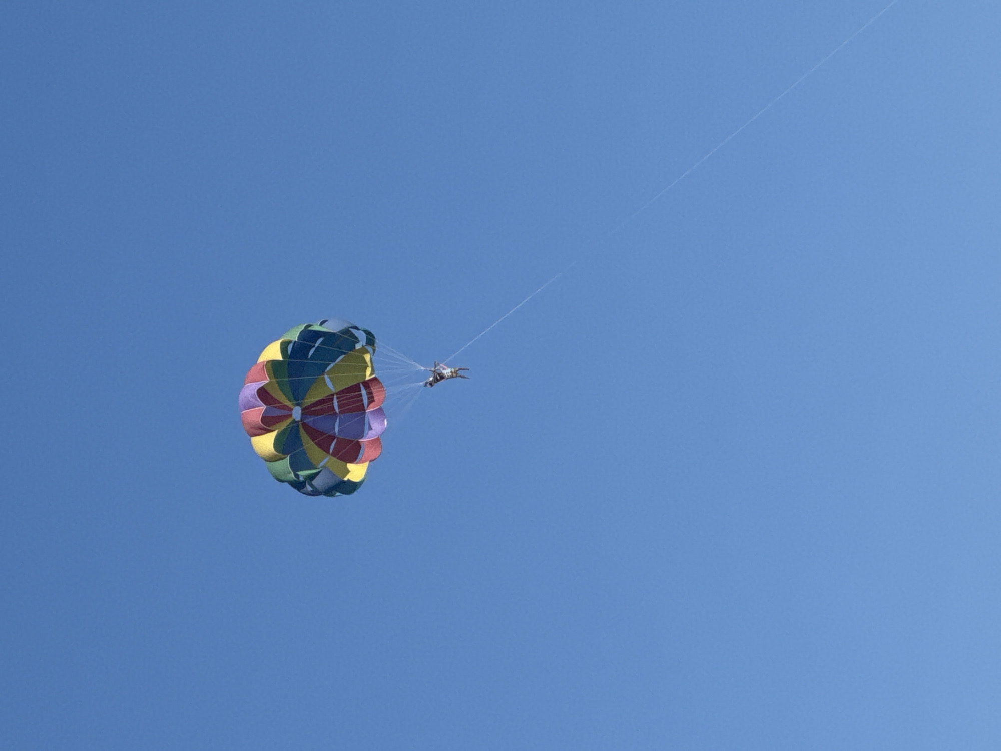 A person is parasailing in a clear blue sky above the city of Sary-Oy, Kyrgyzstan. The parasail is colorful, with sections of yellow, green, blue, purple, and red. The person is suspended below the parasail, with their legs extended and arms up. A thin rope connects the person to a boat that is not visible in the frame. The sky is a bright, cloudless blue.