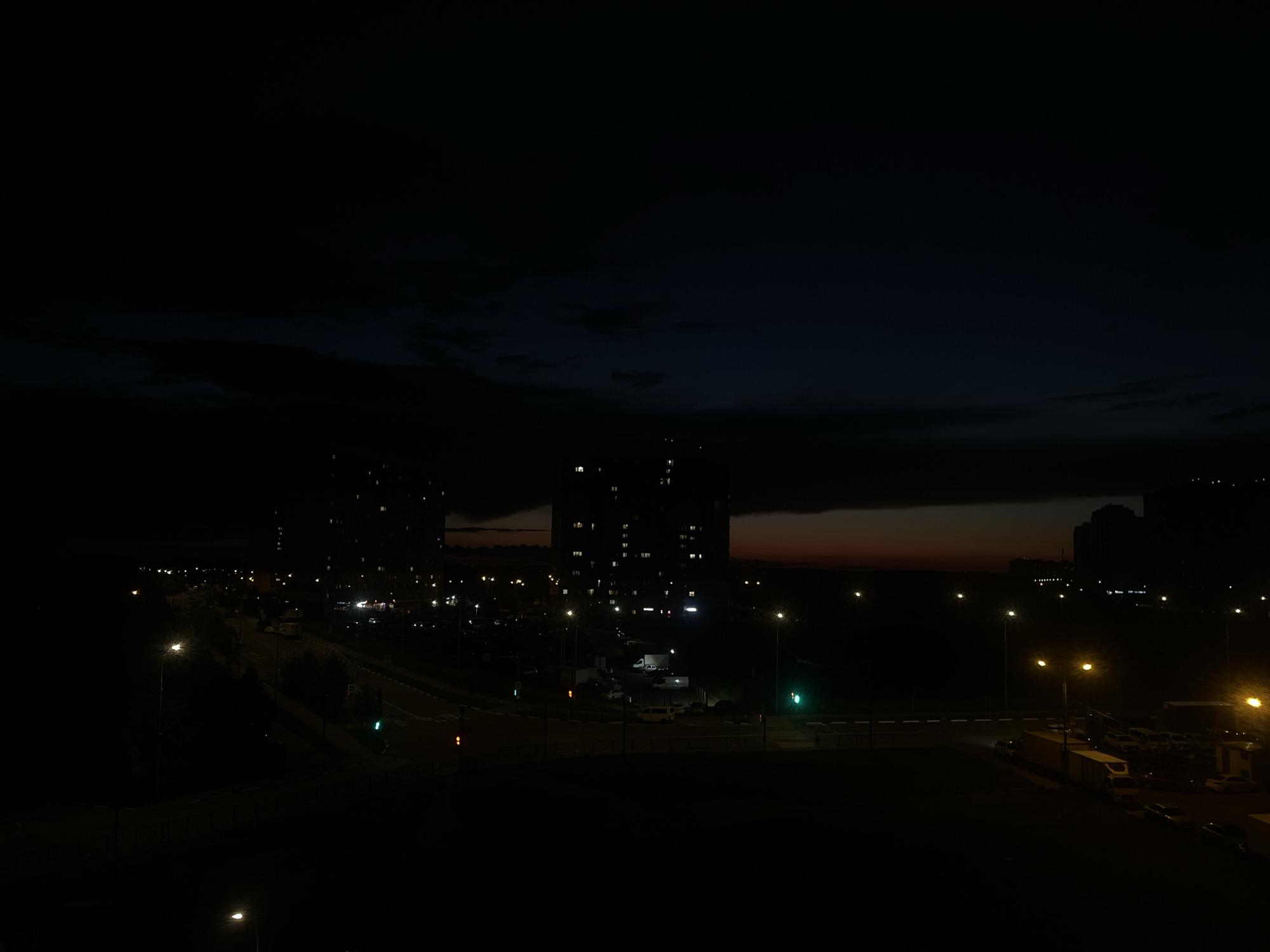 This media file depicts a nighttime urban scene in Domodedovo, Russia. The image is dark, with the sky transitioning from a deep blue to black, suggesting either dusk or pre-dawn hours. Wispy clouds are visible in the upper portion of the frame.
The main subjects are several tall apartment buildings, their windows lit, creating a grid pattern against the dark sky. Below the buildings, a road is visible, illuminated by streetlights and traffic signals. The road appears to be an intersection with at least two traffic lights, one showing green. Several vehicles, including what appear to be cars and a large truck, are present on or near the road, some parked in a lot.
The overall setting is a residential area with infrastructure typical of a suburban or city environment. The presence of illuminated buildings and vehicles indicates human activity, likely during a period of rest or commute. The mood conveyed is one of quietness and the ongoing life of a city at night. There is no visible text in the image.