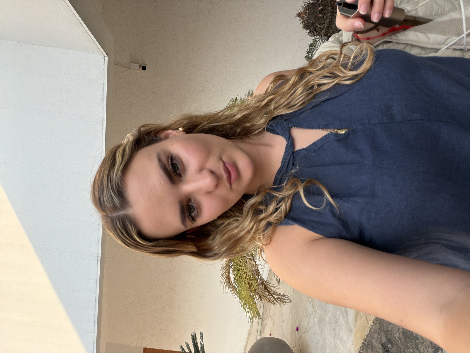 A young adult female is depicted in a close-up, waist-up view, appearing to take a selfie. She is centered in the frame, directly facing the camera with a neutral expression and a slight pout. Her long, wavy hair, blonde with darker roots, is parted centrally. She has green eyes and is wearing a navy blue sleeveless top with a keyhole neckline, accessorized with a gold pendant necklace and a small gold stud earring in her left ear. Her right hand, partially visible on the lower right, holds a dark metallic object alongside a light-colored, possibly beige or off-white, quilted garment draped over her arm.
The background reveals an outdoor, possibly residential, patio or courtyard setting within the city of San Bernardino Tlaxcalancingo, Mexico. The environment features light beige stucco walls. An overhead structure casts a prominent diagonal shadow across a lighter-colored upper wall section. To the left, a large dark gray planter contains a green palm-like plant. Further foliage is partially visible on the far right. The ground is paved with light-toned tiles or concrete, and a few small pink petals are scattered near the planter. A small, dark-colored security camera is mounted on a white base on the upper right beige wall. The scene is lit by natural light, indicating an outdoor daytime setting.