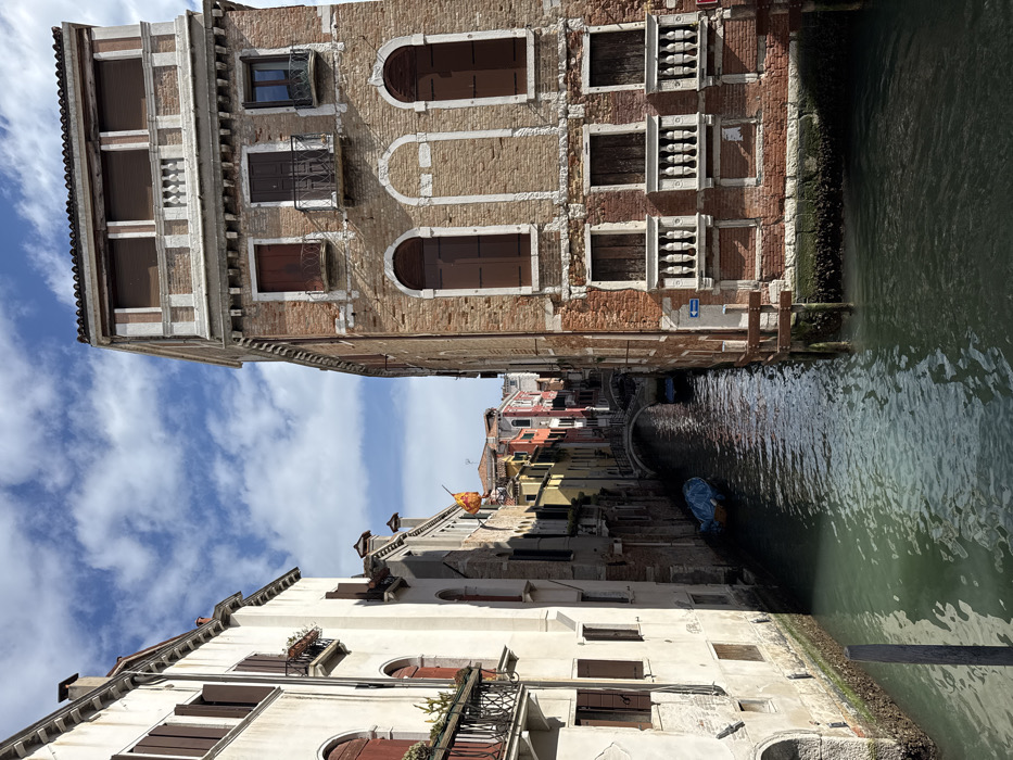 The image displays a narrow canal in Venice, Italy, framed by multi-story buildings under a partly cloudy blue sky.
The building on the left exhibits a cream-colored stucco facade with multiple windows, some fitted with dark brown wooden shutters that are partially open. Balconies with black metal railings, some holding planted greenery, project from its upper levels. The base of this structure shows visible algae growth at the waterline.
The building on the right features a weathered reddish-brown brick facade with exposed mortar. It contains several windows with closed dark brown wooden shutters; some are arched, while lower-level windows are fronted by white stone balustrades. A prominent upper-floor balcony or loggia includes a white balustrade and closed brown roller shutters. A blue directional arrow sign, pointing left, is affixed to the facade near the water level, where extensive algae growth and two wooden mooring posts are present.
The canal water is a murky greenish-brown, reflecting the sky. In the middle distance, a small light blue boat, covered with a blue tarp, is moored along the left bank. Further down the canal, a small arched pedestrian bridge crosses the waterway, beyond which a line of multi-colored buildings (yellow, red, orange) is visible. A yellow and red flag, potentially depicting a winged lion, flies from a building on the left bank in the distance.
No individuals or active watercraft are present in the scene.