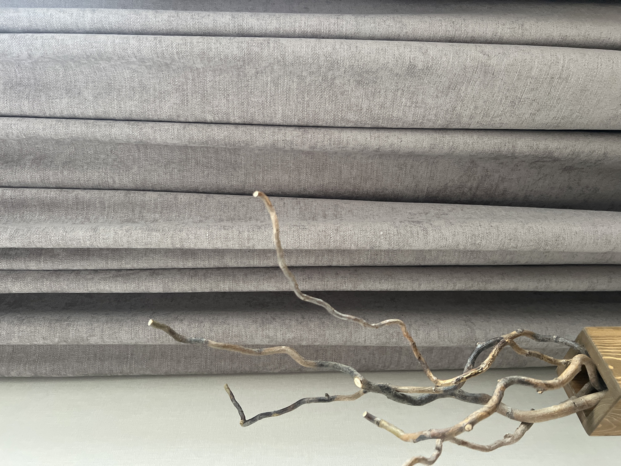A still life image features a decorative arrangement of dried branches in a wooden planter placed against a textured gray curtain. The branches are gnarled and varied in color, ranging from light brown to dark gray. To the left of the branches is a white, textured wall. The scene is likely an indoor setting, possibly a home or commercial space, in Penza, Russia. There are no people or animals visible in the image, and no discernible actions or interactions are taking place. The lighting appears to be natural, coming from an unseen source. The composition is simple and focused on the contrast between the organic forms of the branches and the geometric folds of the curtain.