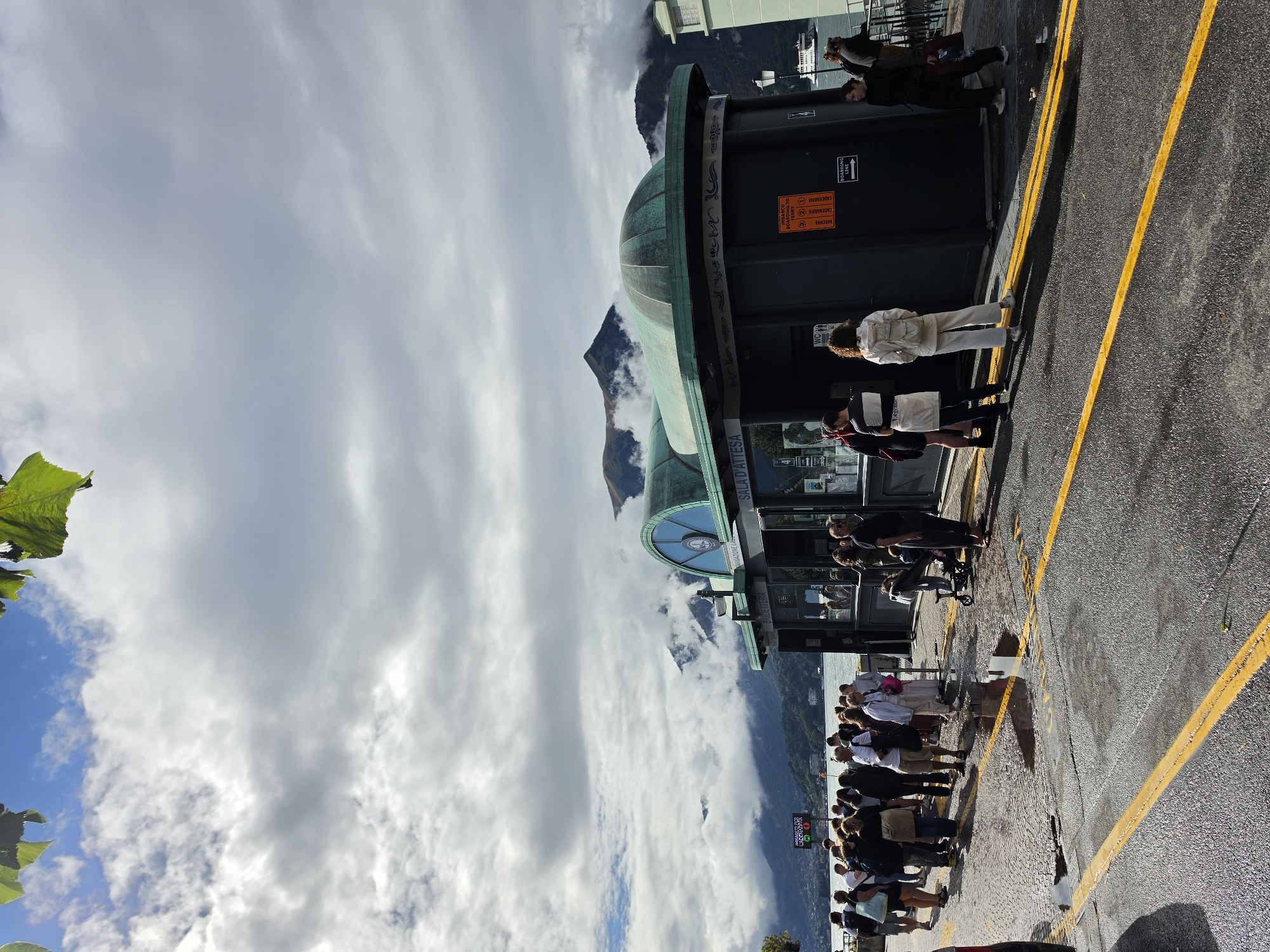 A group of people are standing in line on a wet asphalt surface marked with yellow lines. They are waiting in front of a dark, modern-style building with a curved, green roof. The building appears to be a ticket booth or waiting area, with signage visible. In the background, a body of water stretches out, with mountains and clouds visible behind it. A ferry boat can be seen in the distance on the water. The overall scene suggests a transportation hub in a scenic, possibly mountainous, location, likely a lakeside area in Italy, specifically San Giovanni, given the context provided. The sky is partly cloudy, with patches of blue visible.