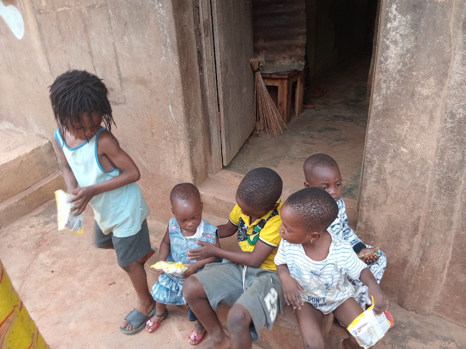 Five young children are gathered outdoors on a concrete stoop next to a weathered building in Shagamu, Nigeria. The children, appearing to be of African descent and ranging from toddler to early primary school age, are engaged in handling small snack packages during the daytime.
On the far left, a child with dreadlocks, wearing a light blue tank top and dark shorts, stands holding a small, white and yellow snack package. To their right, a younger child in a blue denim-style dress is seated, also holding a similar package. Three other children are seated closely together on the stoop to the right. One child wears a yellow and black patterned t-shirt and shorts, looking towards the others. Another child, wearing a white and blue striped t-shirt with partial text "DON'T G_ _ ME" visible on it, holds a snack package. Behind them, another child is partially visible in a light-colored patterned top.
The setting features a textured, brownish-orange wall and a dark wooden doorway leading into a dimly lit interior. Inside the doorway, a wooden stool and a broom made of natural fibers are visible. The children are either barefoot or wearing simple sandals. The lighting suggests it is broad daylight, likely mid-morning or afternoon. The children appear calm and focused on the snacks.