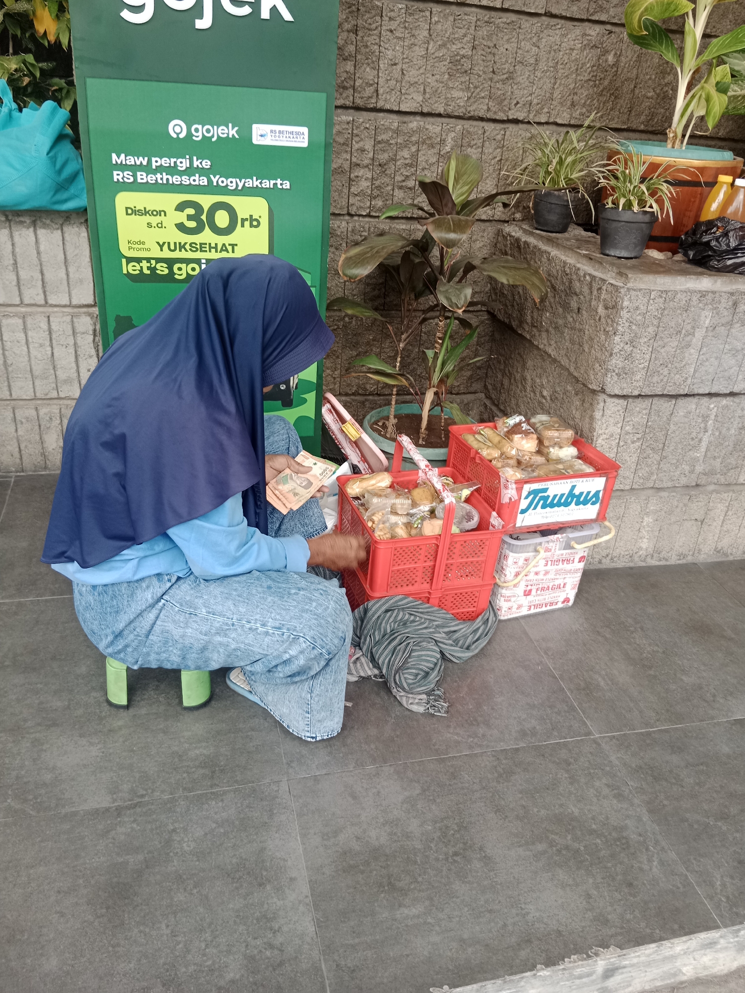 In Yogyakarta, Indonesia, a woman wearing a navy blue hijab and light blue jeans sits on a small green stool, counting a wad of Indonesian Rupiah bills. She is surrounded by red plastic baskets filled with baked goods. One basket sits in front of her, and another is stacked on top. A grey and white cooler bag labeled "FRAGILE" is placed beneath the stacked baskets. Behind the woman, a green banner promotes Gojek rides to RS Bethesda Yogyakarta with a discount code. The scene takes place on a grey tiled floor, with a textured stone wall in the background. Potted plants are visible on a ledge behind the baskets.