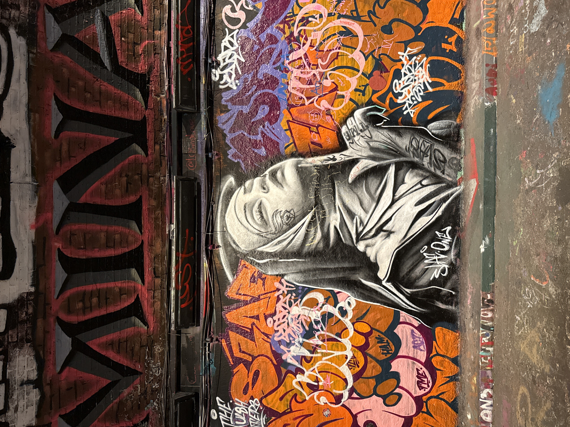 The image depicts a wall in London, United Kingdom, covered in graffiti. A grayscale mural of a person with their head covered is the focal point, surrounded by colorful abstract graffiti patterns in orange, pink, and purple hues. To the left of the mural, a vertical section features larger block letters in red and black. Exposed pipes and wiring run along the wall's surface. The rightmost section shows a rough, textured wall with faded paint and markings.