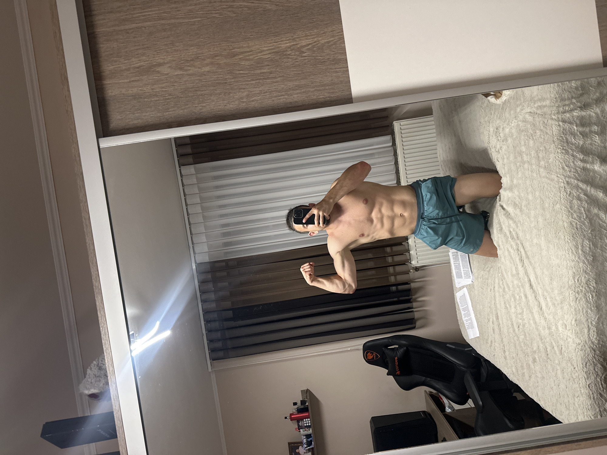 An indoor, vertical mirror selfie captures a male subject in what appears to be a bedroom located in the city of Saraia, Bulgaria.
The subject, a male with dark hair tied back in a bun and a beard, is shirtless, exposing a muscular torso with defined abdominal muscles. He is wearing teal-colored athletic shorts and is kneeling on a light-colored, fluffy bedspread. The subject is flexing his right bicep and holding a black smartphone directly in front of his face, obscuring it.
The room is reflected in a large, floor-to-ceiling mirror, likely part of a sliding wardrobe, which occupies the right half of the image. The wardrobe features a light wooden panel with a grain texture on the right side and a white frame with a silver sliding track. Above the wardrobe, a black electronic device and a white, fluffy object are visible on a ledge, beneath a light-colored ceiling with white crown molding.
To the left of the subject, also reflected in the mirror, is a beige-colored wall. A window is covered by white sheer curtains and dark brown opaque curtains. Below the window, a white radiator is installed. Further to the left, a black gaming chair with red accents and a "COUGAR" logo is positioned next to a dark-colored desk or table, on which a black speaker or computer tower rests. Above the desk, a wall-mounted shelf displays several items, including framed photographs, a red container, and a black bottle. Two sheets of paper with printed text are visible on the bedspread near the subject's left knee. The room is illuminated by artificial light, causing a strong glare to be reflected in the upper center of the mirror.