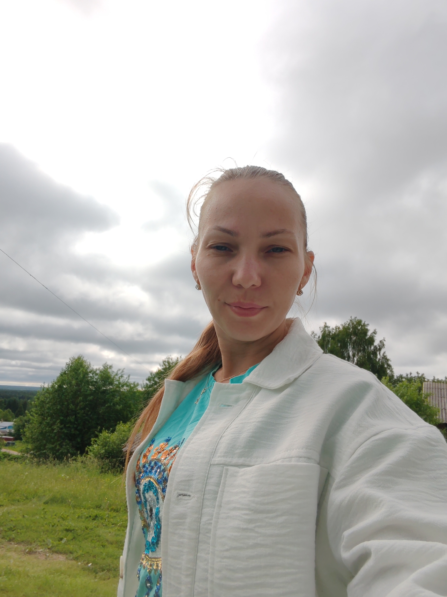 A medium shot selfie shows a fair-skinned woman with blonde hair pulled back and some strands loose around her face, looking directly at the camera. She is wearing a light blue top with intricate gold and blue patterns and a loose white jacket. She has earrings in her ears and a necklace. She stands outdoors with a blurred background of green trees, a grey sky with clouds, and a hint of a house with a grey roof on the right. A thin black wire runs diagonally across the image from the left. The overall scene suggests an overcast day in a rural or semi-rural setting, potentially in the city of Chukhlom, Russia.
