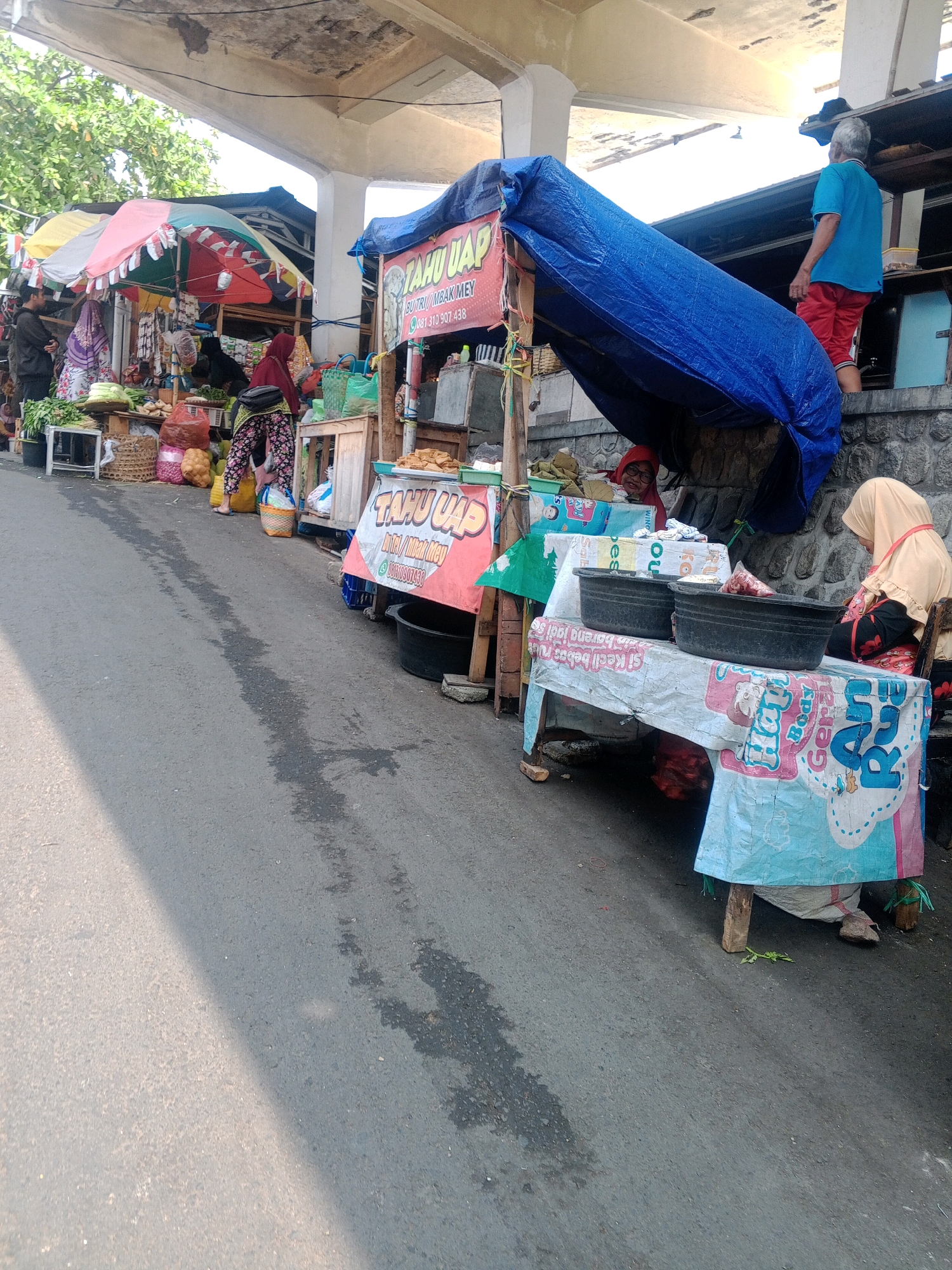 This image depicts a street-side market scene in Wonogiri, Indonesia. The market is situated alongside an asphalt road, with a structure providing shade and shelter overhead. Various stalls are set up, displaying a range of goods.
In the foreground, the road has patches of wetness and is partially illuminated by sunlight. To the right, a market stall is covered with a blue tarp, with a banner that reads "TAHU UAP" (steamed tofu) and a phone number. Underneath the tarp, a person in a red headscarf is visible. Next to this stall, another stall is covered with a white and blue patterned tarp, displaying two black plastic basins. A woman wearing a light yellow hijab is seated at this stall.
Further to the left, several other market stalls are visible. One stall has multiple colorful umbrellas providing shade and displays produce such as vegetables and fruits. People can be seen browsing and working at these stalls. Bags of goods, baskets, and various food items are arranged for sale. A man in a blue shirt and red shorts is standing near the stalls on the right side of the image, facing away from the camera and looking towards the overhead structure. The environment appears to be an outdoor market with a mix of temporary structures and natural elements like trees in the background.