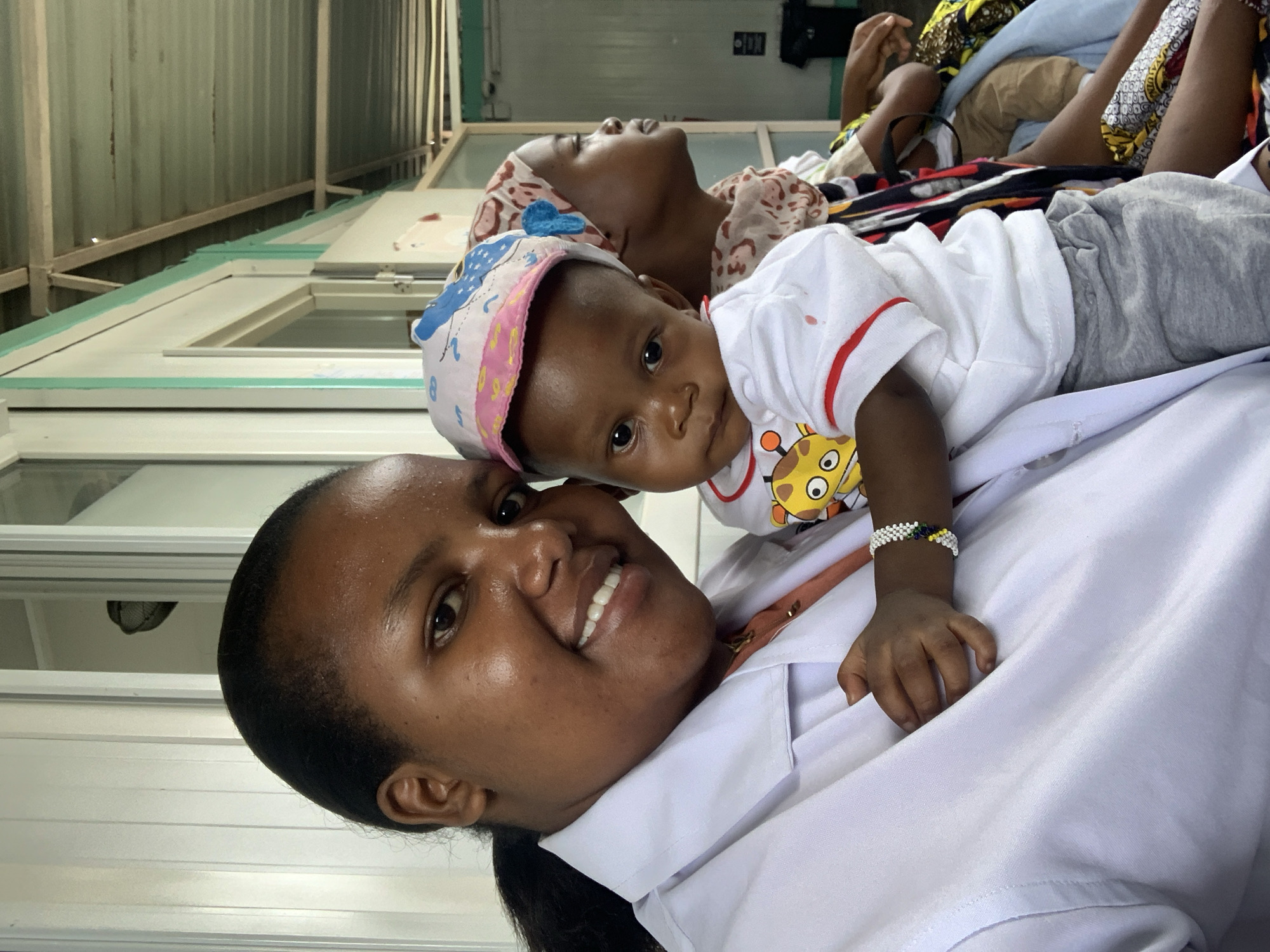 A mid-shot photograph captures an adult woman holding an infant in a waiting area located in Dar es-Salaam, Tanzania. The primary subject, a woman with dark skin and dark hair in a ponytail, wears a white lab coat over a light peach top and a thin gold chain. She smiles and looks directly at the camera. She holds an infant, also with dark skin, who wears a white and pink patterned hat, a white t-shirt with red sleeve trim and a yellow giraffe graphic, and gray shorts. A white, green, yellow, and blue beaded bracelet is on the infant's left wrist. The infant looks towards the camera with a neutral expression. Behind the infant and to the right, a second adult woman is partially visible, seated and looking in profile to her right. She wears a patterned headscarf and a dark garment. The background environment consists of white paneled walls or doors with light green trim, and an exposed corrugated metal roof supported by wooden rafters. A dark waste bin is visible in the distant background against a light wall.