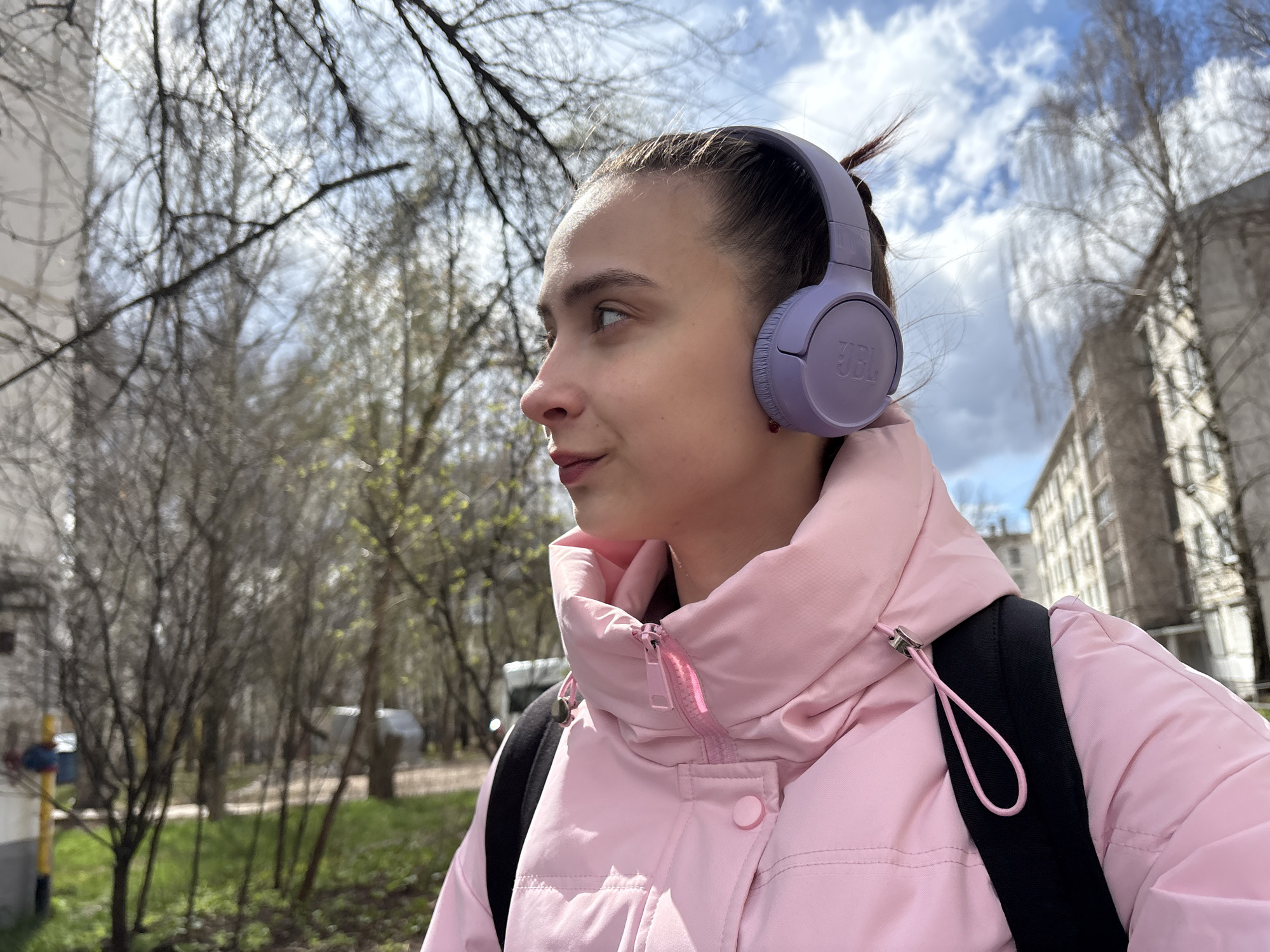 The image features a young woman outdoors in a city or town, likely Chaikovsky, Russia, during early spring. She is depicted from the chest up, in profile, looking towards the right side of the frame with a calm, observant expression. Her dark hair is pulled back, and she is wearing lavender over-ear headphones with the brand "JBL" visible on the right earcup. She is dressed in a light pink puffy jacket with a high collar and a pink zipper. A black strap from a backpack is visible over her right shoulder.
The background consists of numerous deciduous trees, many still bare, but some showing the first signs of budding green leaves, strongly indicating an early spring season. The sky is bright with scattered white clouds, suggesting a partly cloudy day. In the distance on the far right, several multi-story buildings, possibly apartment blocks, are visible, contributing to the urban or suburban setting. The lighting suggests it is daytime. The woman appears to be engaged in a casual activity, such as walking or commuting, while listening to audio.