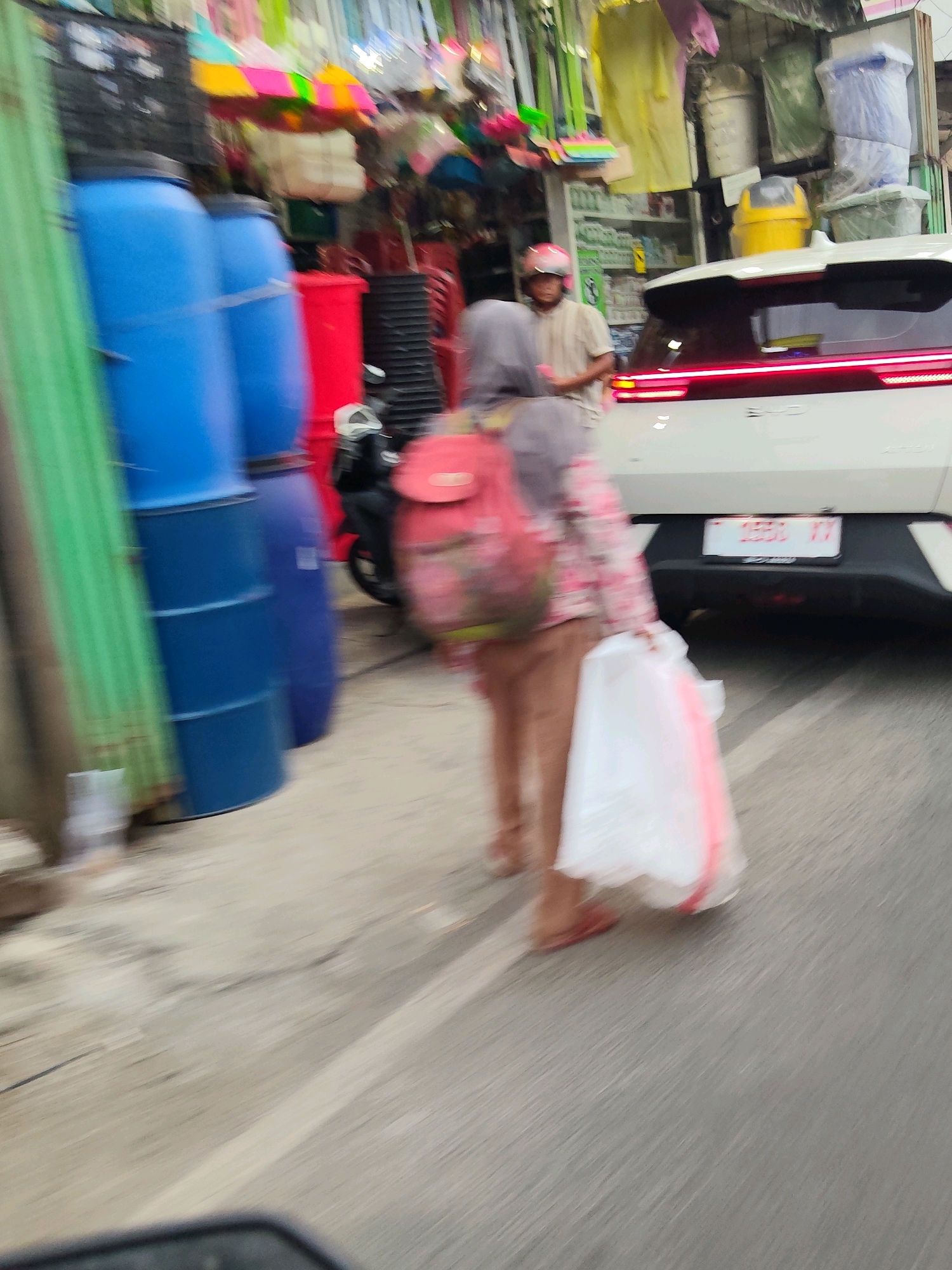 The image captures a street scene in Purwakarta, Indonesia, likely a market or roadside stall area. In the foreground, two individuals are blurred due to motion. The person on the left, wearing a gray headscarf and a patterned shirt, is carrying a red backpack and a white plastic bag. The person behind them, wearing a light-colored shirt and a helmet, appears to be a motorcyclist. To the left, several large blue barrels and a red plastic container are visible, along with some green vertical objects, possibly a fence or covering. In the background, a white car with a license plate is parked. The environment suggests an outdoor setting with various goods displayed, typical of a bustling market.