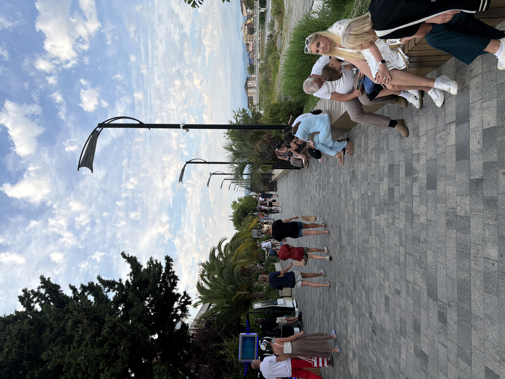 The image depicts a lively outdoor scene in Sochi, Russia, likely during the daytime given the bright sky. A group of people are enjoying a leisurely stroll along a paved promenade lined with palm trees and lampposts. The sky is a soft blue, dotted with wispy white clouds. In the foreground, a family or group of friends are gathered on a bench, with a woman in a white lace dress and a child being held by an older man. Another woman in a white top and sunglasses is seated next to them, looking at the camera.
Further down the promenade, several individuals are walking in the same direction. Some are dressed casually in t-shirts and shorts, while others wear dresses or skirts. One person is holding a brown paper bag. There is a kiosk with a screen and some seating, suggesting a public area or amenity. In the background, buildings and greenery are visible, hinting at a developed urban or resort area. The overall mood appears relaxed and social, with people enjoying the pleasant weather and surroundings. The pavement is made of grey interlocking stones.