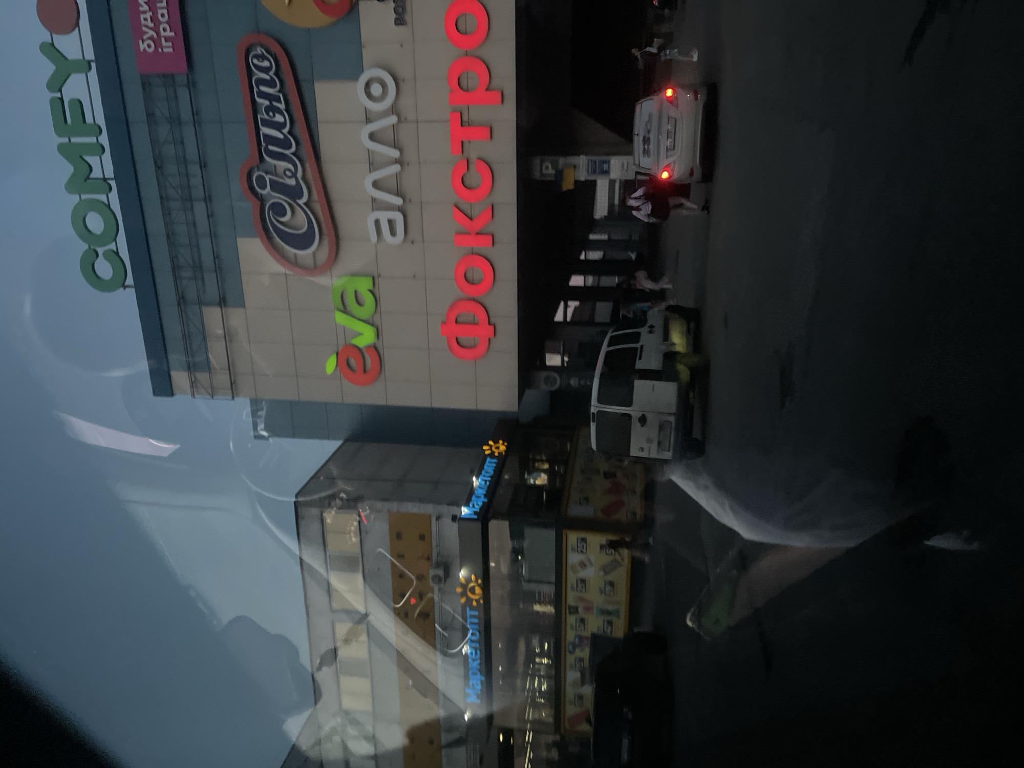 The image shows a street scene in front of a shopping center. The shopping center has several prominent signs, including "COMFY," "Єва," "алло," "Сіпьно," and "ФОКСТРОТ". There are multiple vehicles visible, including a white van, a white sedan with its brake lights illuminated, and a black car. Several people are seen in the image, some walking on the pavement and others near the vehicles. The lighting suggests it is dusk or evening. Reflections are visible on the surface from which the photograph was taken, obscuring parts of the scene.