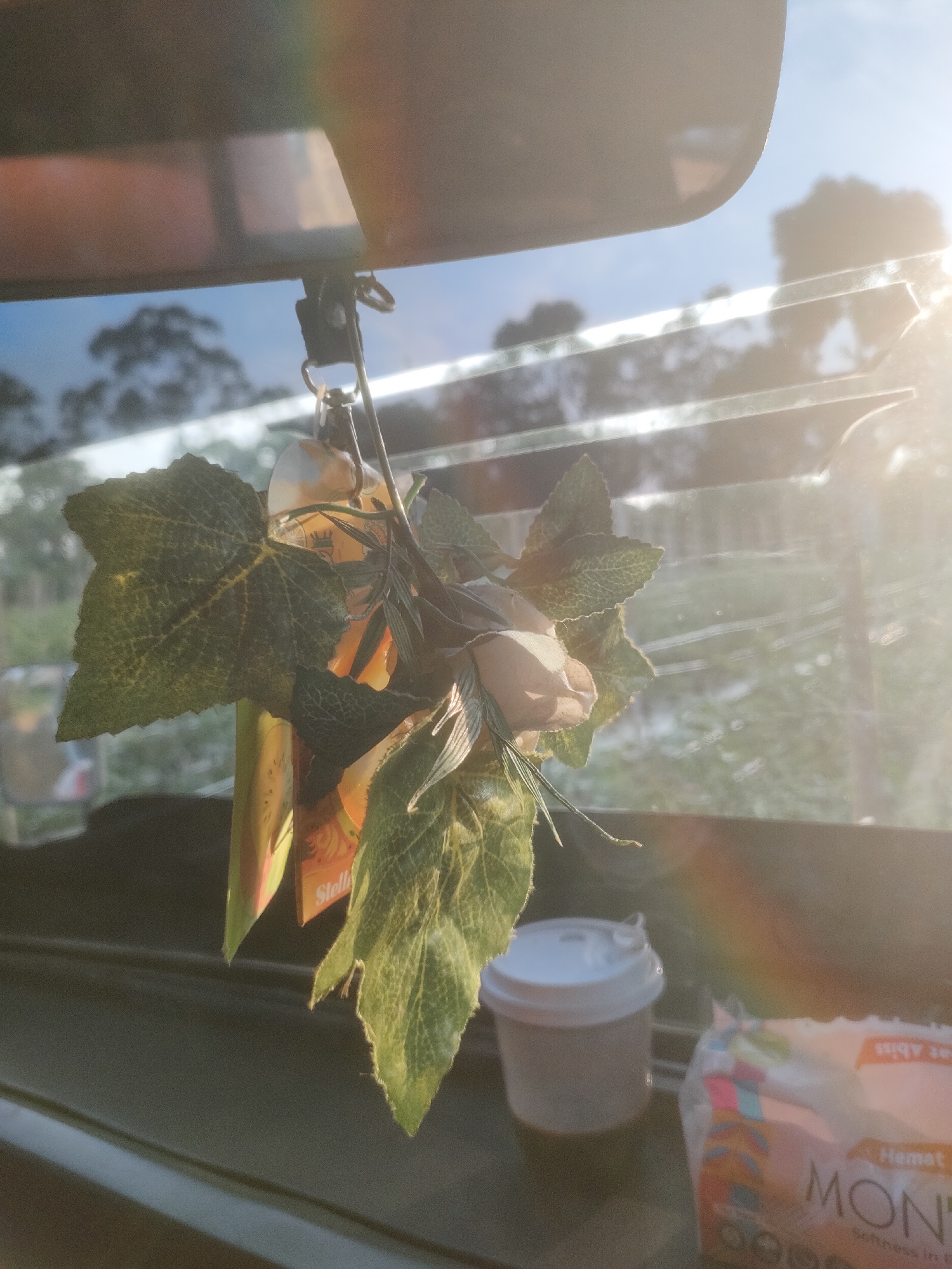 The image depicts a close-up view of a car's interior, specifically the area around the rearview mirror. A decorative item, consisting of artificial leaves and a small fabric flower, hangs from the mirror. To the right of this decoration, a small white paper cup is visible, possibly a disposable coffee cup. Further to the right, a package of wet wipes is partially visible, with the brand name "MON..." and text that appears to say "Hemmat SOFTNESS IN P" discernable. The background through the car's windows shows a bright, sunny outdoor scene with trees and a blue sky, indicating daytime. A rainbow-like light refraction is present, suggesting strong sunlight. The specific location of Padang Dalom, Indonesia, is not visually evident from the image itself.