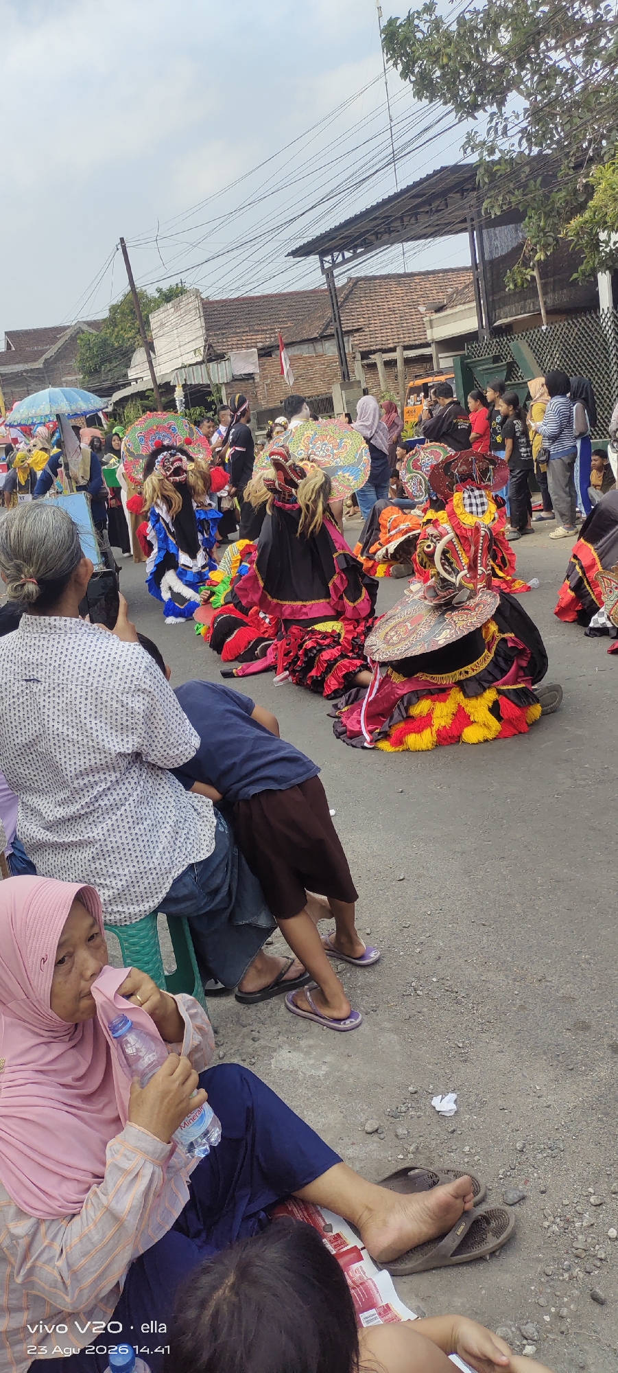 The image captures a lively street scene, likely a festival or parade, in Simo, Indonesia. In the foreground, a woman wearing a pink hijab and striped long-sleeved shirt sits on the ground, holding a bottle of water. Next to her, a person in a blue t-shirt and brown shorts is leaning against someone seated on a green stool, who is wearing a white patterned shirt.
The main focus of the scene is a group of performers in elaborate, colorful costumes, resembling traditional Javanese dance. They are wearing masks that depict animalistic or demonic faces, with large, vibrant headdresses and layered, ruffled skirts in red, yellow, and black. Some performers are in motion, while others appear to be resting or interacting with the crowd. The performers are on a paved road, which is lined with spectators.
In the background, more people are visible, some standing and watching the performance, while others are seated. Buildings with terracotta tile roofs are visible along the street, suggesting a residential or commercial area. Overhead, a tangle of electrical wires crisscrosses the sky, typical of urban environments in Indonesia. An umbrella, possibly for shade or decoration, is also visible in the crowd. The overall atmosphere appears to be festive and engaging, with a clear cultural element from the performers' attire.