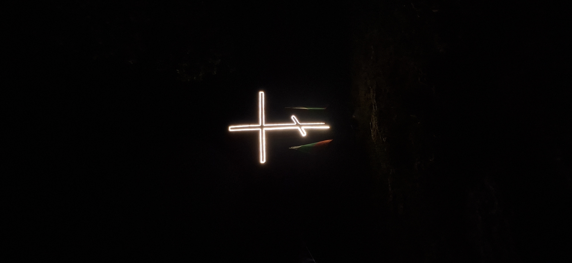 An outdoor nighttime photograph displays a prominently illuminated Eastern Orthodox cross, constructed from bright, white-yellow light strips, centrally positioned against a completely dark background. The cross features a main horizontal bar, a shorter horizontal bar positioned above the main one, and a lower diagonal bar slanted downwards from upper left to lower right. To the immediate left of the cross, a partially unfurled flag with visible stripes of green, white, and red is discernible, consistent with the national flag of Bulgaria, catching some light from the cross. To the immediate right, another flag is present, primarily exhibiting a vertical green stripe. The ground directly beneath the cross and flags is dark and uneven, suggesting natural, unpaved terrain. In the upper right background, faint, dark silhouettes of trees or foliage are barely visible against the dark sky. The scene is located in the city of Lyaskovets, Bulgaria.
