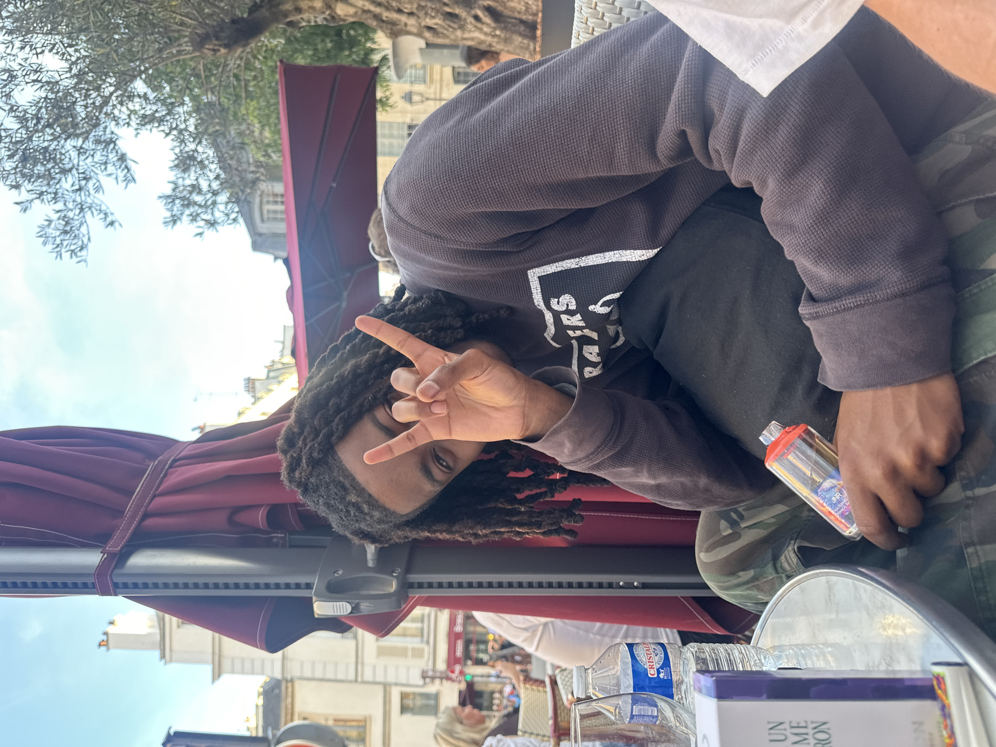 A person with dreadlocks is seated at an outdoor cafe in Paris, France, under a red umbrella. The person is wearing a dark hoodie and camouflage pants, making a peace sign with their left hand. In their right hand, they hold a clear, rectangular object. On the table are bottles of water, a glass, and a book titled "Un M... Ron". The background shows buildings characteristic of Parisian architecture. The photo appears to have been taken on a sunny day.