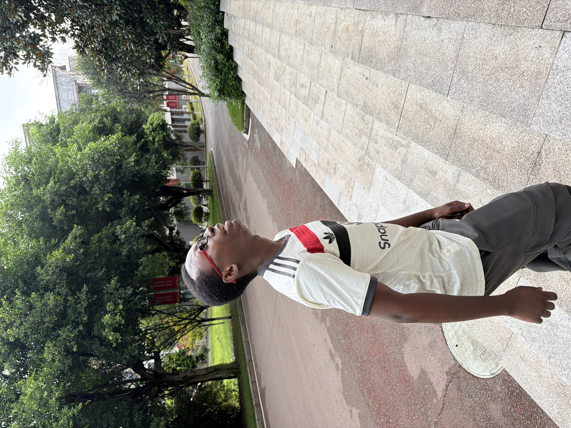 In Jiangjin District, China, a young Black male with short, dark hair and glasses is captured from the chest up, looking upwards. He is wearing a white t-shirt with black and red stripes across the chest, featuring a prominent Adidas logo and text that appears to read "Snap". He is also wearing grey cargo pants. The man is positioned on a light-colored stone staircase, with his left arm bent at the elbow and his hand relaxed at his side. To his left, a road with a reddish-brown surface is visible, bordered by green grass and trees. In the background, a building with a light-colored facade and a gray roof stands amidst lush green trees and foliage, suggesting an outdoor, possibly campus or park setting. The sky is overcast, contributing to a diffused lighting condition.