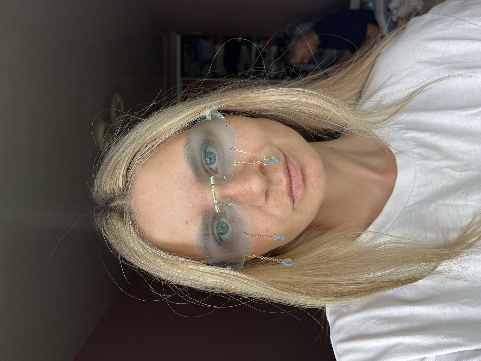 A fair-skinned blonde woman with light blue eyes is centered in a slightly rotated, close-up, high-angle shot. She is wearing unusual eyewear consisting of clear, vaguely flower-shaped frames with gold-colored nose pieces and delicate gold chains hanging from the bottom corners, each ending in a small, light blue bead. She is wearing a white t-shirt. The background is divided between a dark brown wall on the left and a blurred area that suggests an interior space with a person in a blue shirt. There is no verifiable evidence in the image to confirm the location as the city of Chaikovsky, Russia.