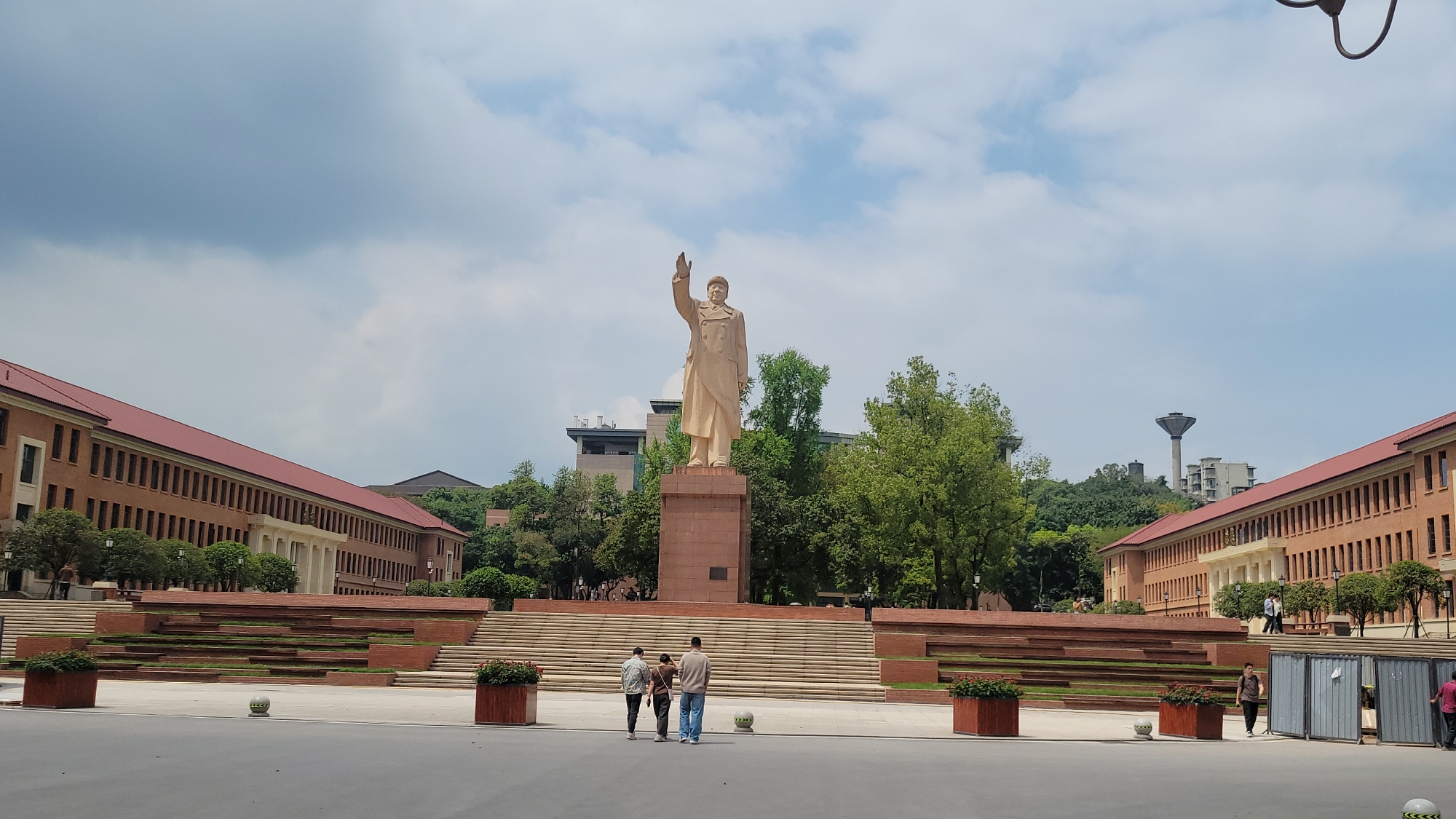 Here is a detailed description of the image:
**Scene:** The image depicts a large public square dominated by a towering statue of Mao Zedong. The statue is placed atop a multi-tiered base constructed of red brick or stone. The statue itself is also made of a light-colored stone. Three people stand facing the statue on the square, which is devoid of other visible activity.
**Architecture:** The square is flanked by two large brick buildings with red roofs, positioned symmetrically on either side of the statue. These buildings feature numerous windows and a classical architectural style. A large tiered staircase leads from the square to the base of the statue, adding to the grandeur of the scene.
**Environment:** The scene is set on a partly cloudy day. Trees and other vegetation surround the square. Some taller structures, possibly water towers or other industrial buildings, can be seen on a hill in the background.
**People:** The three individuals standing in the square appear to be tourists or visitors. Their posture suggests they are admiring the statue.
**Objects:** In addition to the statue, buildings, and trees, there are flower boxes arranged along the base of the steps and some temporary-looking barriers on the right.
**Actions/Interactions:** The primary action is the observation of the statue by the three individuals.
**Location:** Given the presence of the statue and the architectural style, it is most likely located in China. Without additional information, it is not possible to determine the exact location.