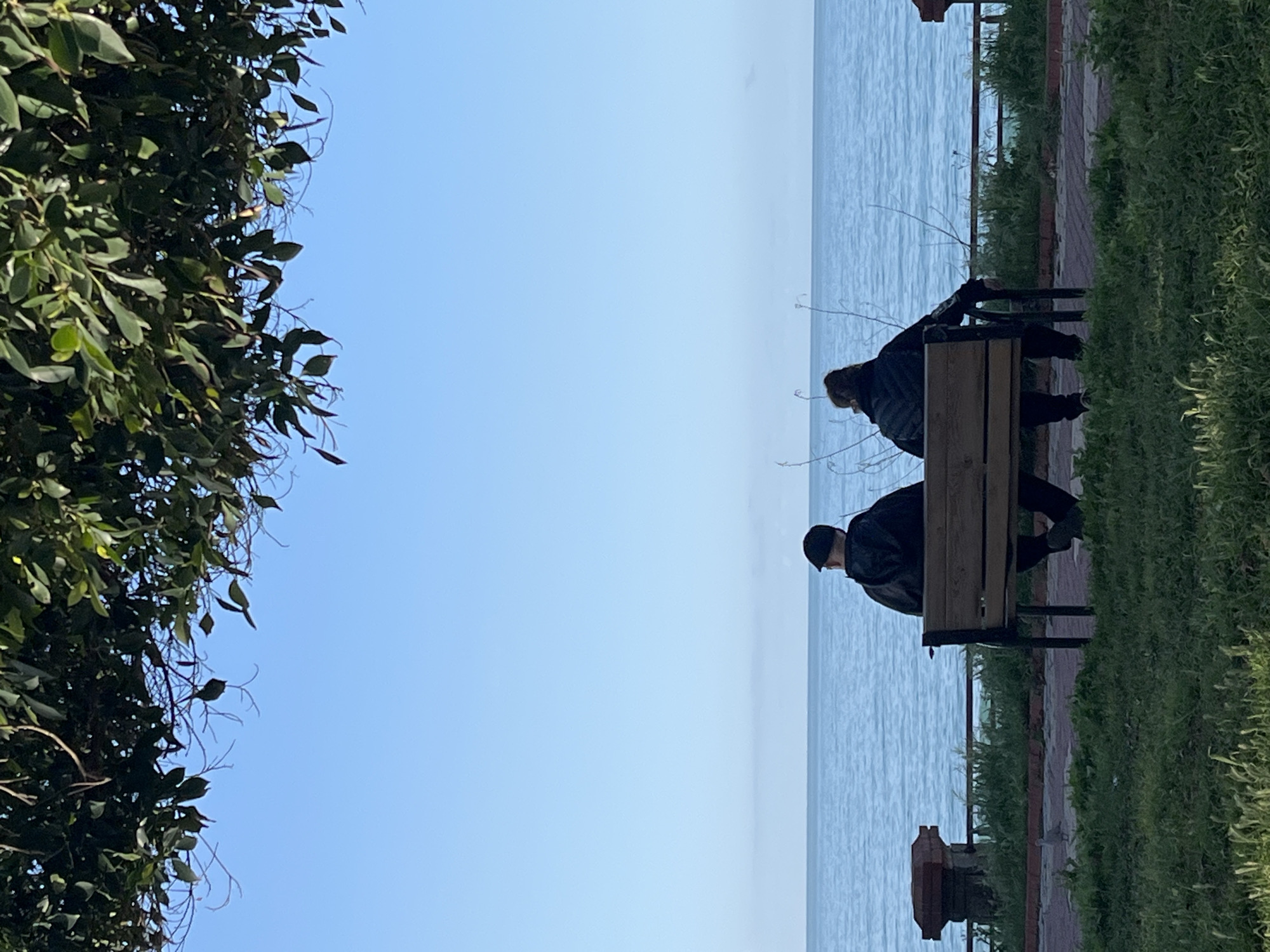 Two people are sitting on a bench facing the sea. The environment includes foliage on one side and a paved walkway with grass on the other. The location is Alanya, Turkey.