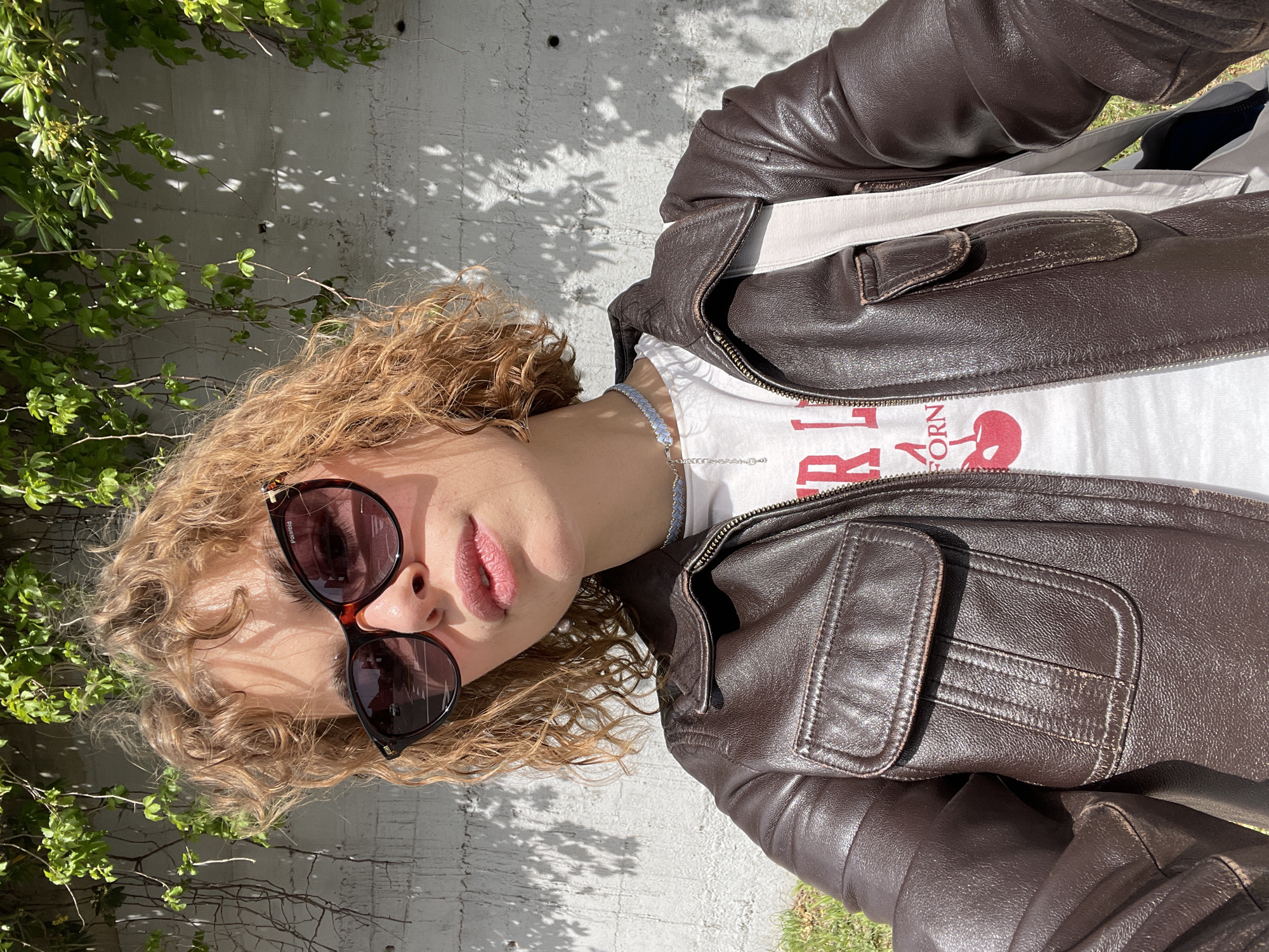 A person with curly blonde hair is wearing sunglasses and a brown leather jacket over a white graphic t-shirt. They are wearing a light blue necklace. The person is positioned against a white wall covered in shadows of leaves and vines. Green vegetation is visible to the left and bottom right. The exact location in Rafailovići, Montenegro, cannot be determined from the provided image.