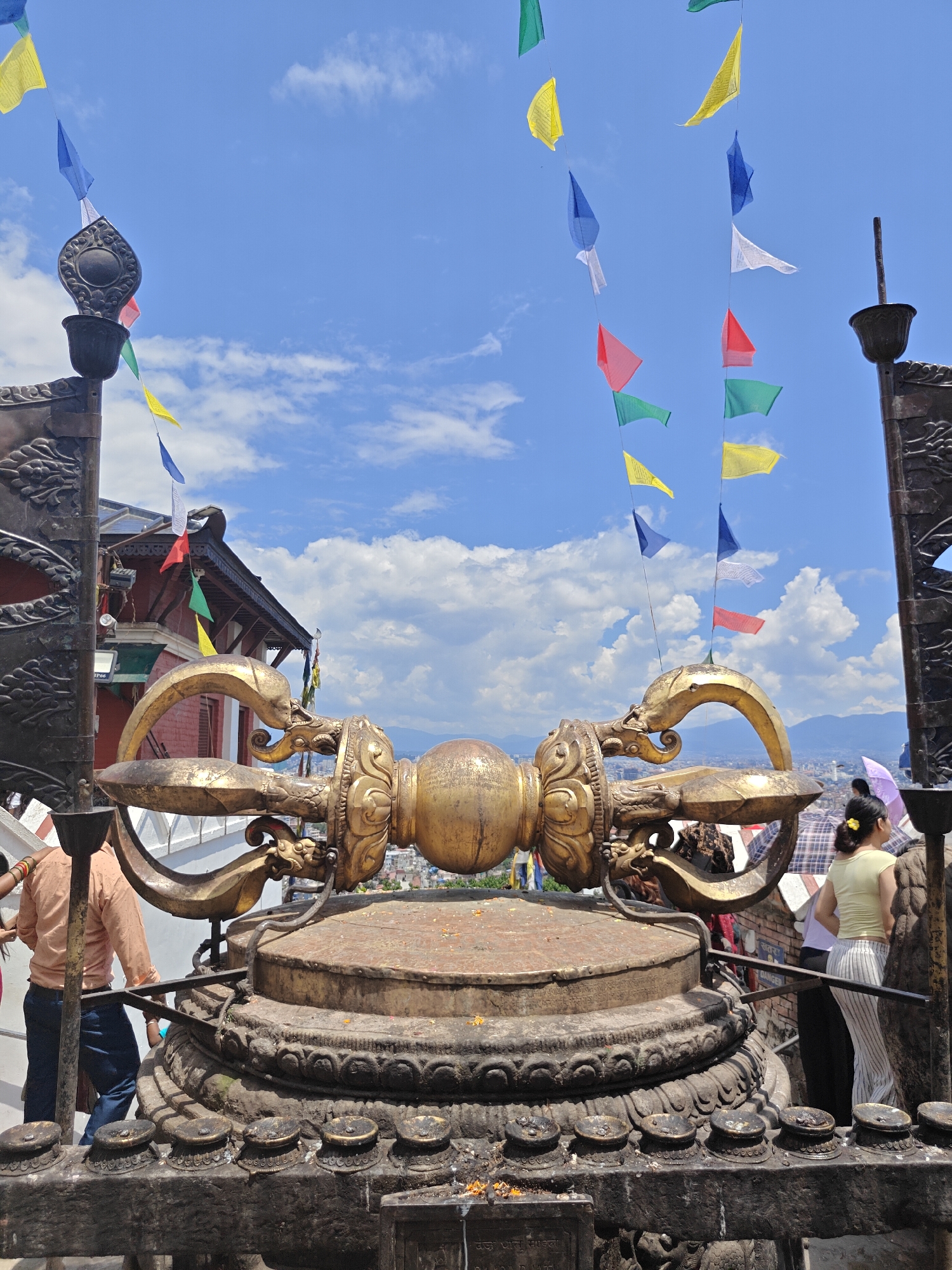 The image captures a golden vajra statue at the Swayambhunath Stupa, also known as the Monkey Temple, located in Kathmandu Metropolitan City, Nepal. The statue is ornate and sits atop a large, circular pedestal adorned with decorative carvings. Prayer flags in various colors – blue, yellow, white, red, and green – are strung across the sky, fluttering in the wind against a backdrop of a clear blue sky with scattered white clouds. Several people are visible in the scene. On the left, a person wearing an orange shirt and blue jeans is standing near the base of a decorative metal structure. To the right, a woman wearing a yellow top and striped pants is seen looking away from the camera, holding an umbrella. Other individuals are partially visible in the background. The environment suggests an outdoor religious site with a view of distant hills or mountains.