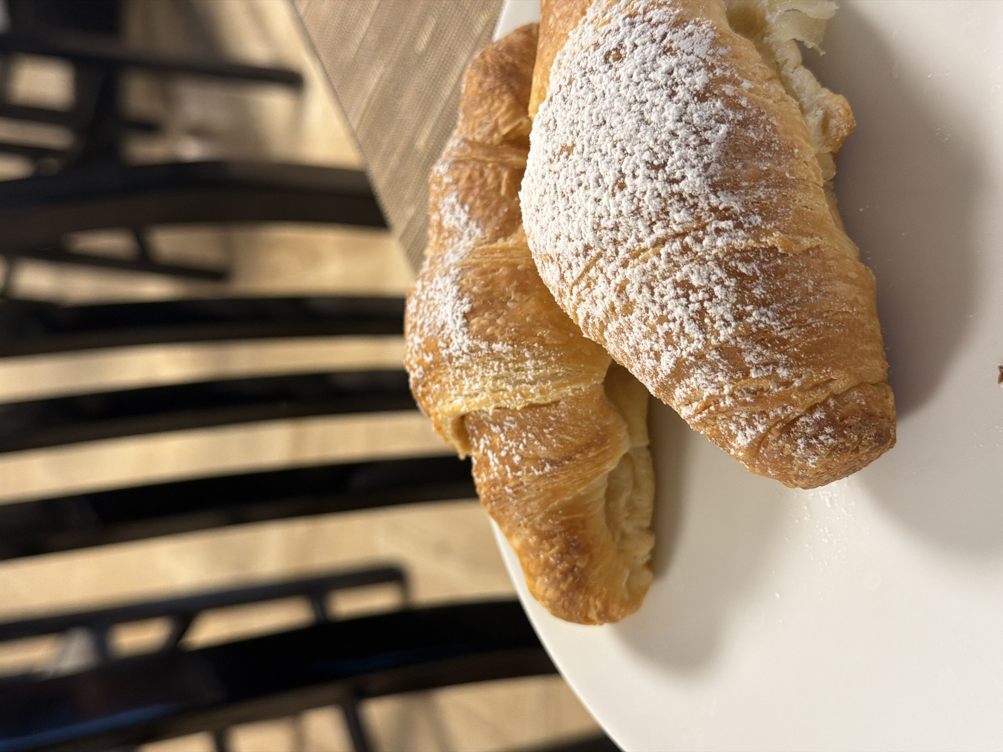 Two croissants are on a white plate. One croissant has powdered sugar on top. The plate is on a table next to a chair with black metal legs. The location is implied to be Trescore Balneario, Italy.