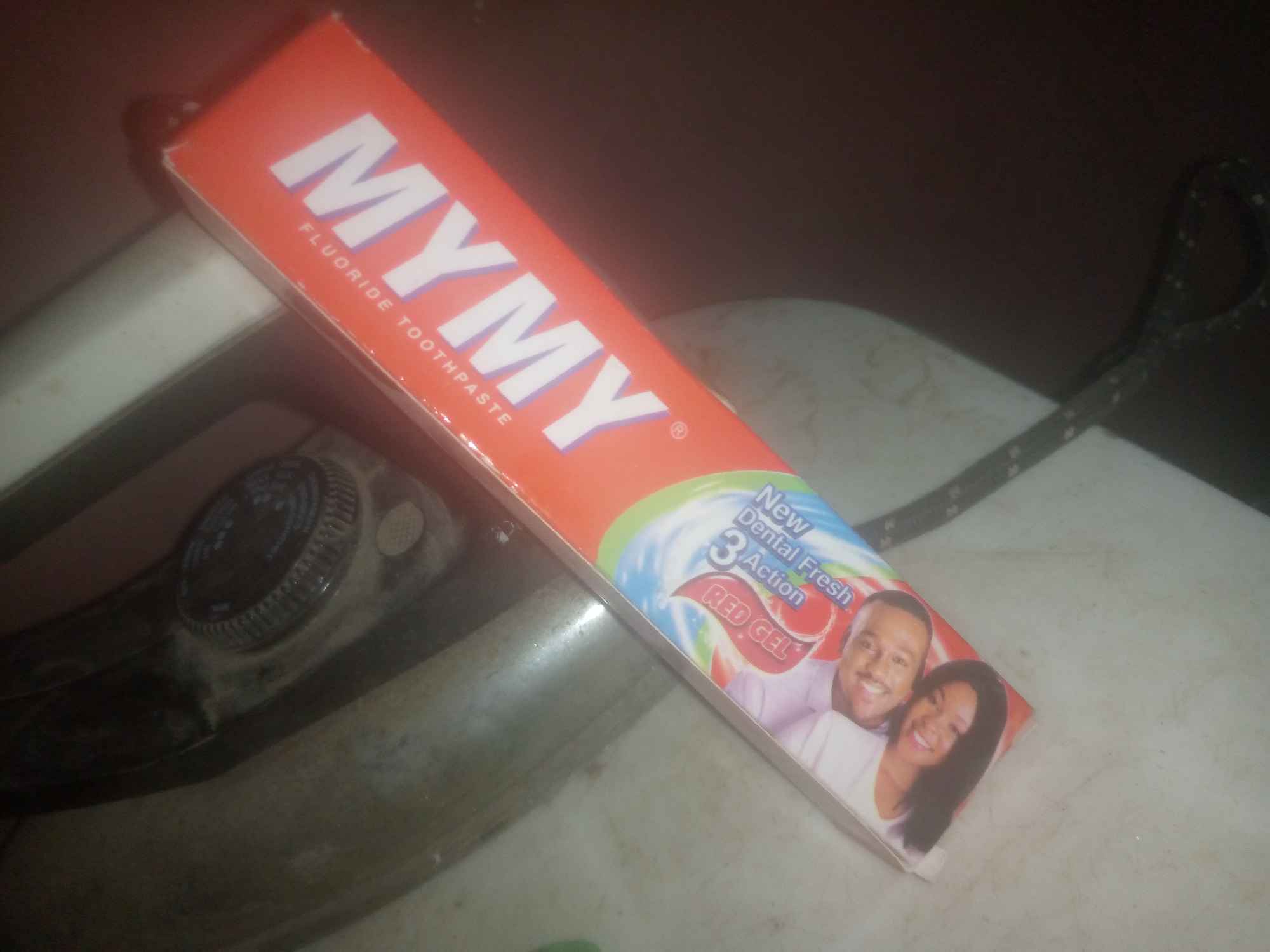 An angled, medium shot shows a tube of "MYMY FLUORIDE TOOTHPASTE" with a bright red and orange color scheme, lying on a light-colored, slightly textured surface. The tube is positioned diagonally, with the top left corner closest to the viewer. The branding "MYMY" is in large, white, capital letters with a light purple outline. Below it, "FLUORIDE TOOTHPASTE" is written in smaller, white, capital letters.
Further down the tube, colorful graphics depict a swirling design of blue, green, and white, with the text "New Dental Fresh 3 Action" in white and blue. Below this, "RED GEL" is written in white, capital letters within a red oval. The bottom right of the tube features a picture of a smiling man and woman. The man, with short dark hair and a light purple shirt, is positioned behind and to the left of the woman. The woman, with long dark hair and a white shirt, is smiling and positioned to the right and slightly in front of the man.
To the left of the toothpaste tube, part of a metallic, grey object with a black dial is visible. The dial has white markings and appears to be part of an appliance, possibly an iron, given its shape. A black power cord with a white pattern is visible in the upper right quadrant of the image, extending out of frame. The overall environment appears to be indoors, possibly a kitchen or laundry area, with subdued lighting. The surface on which the objects rest is speckled with small dark marks.