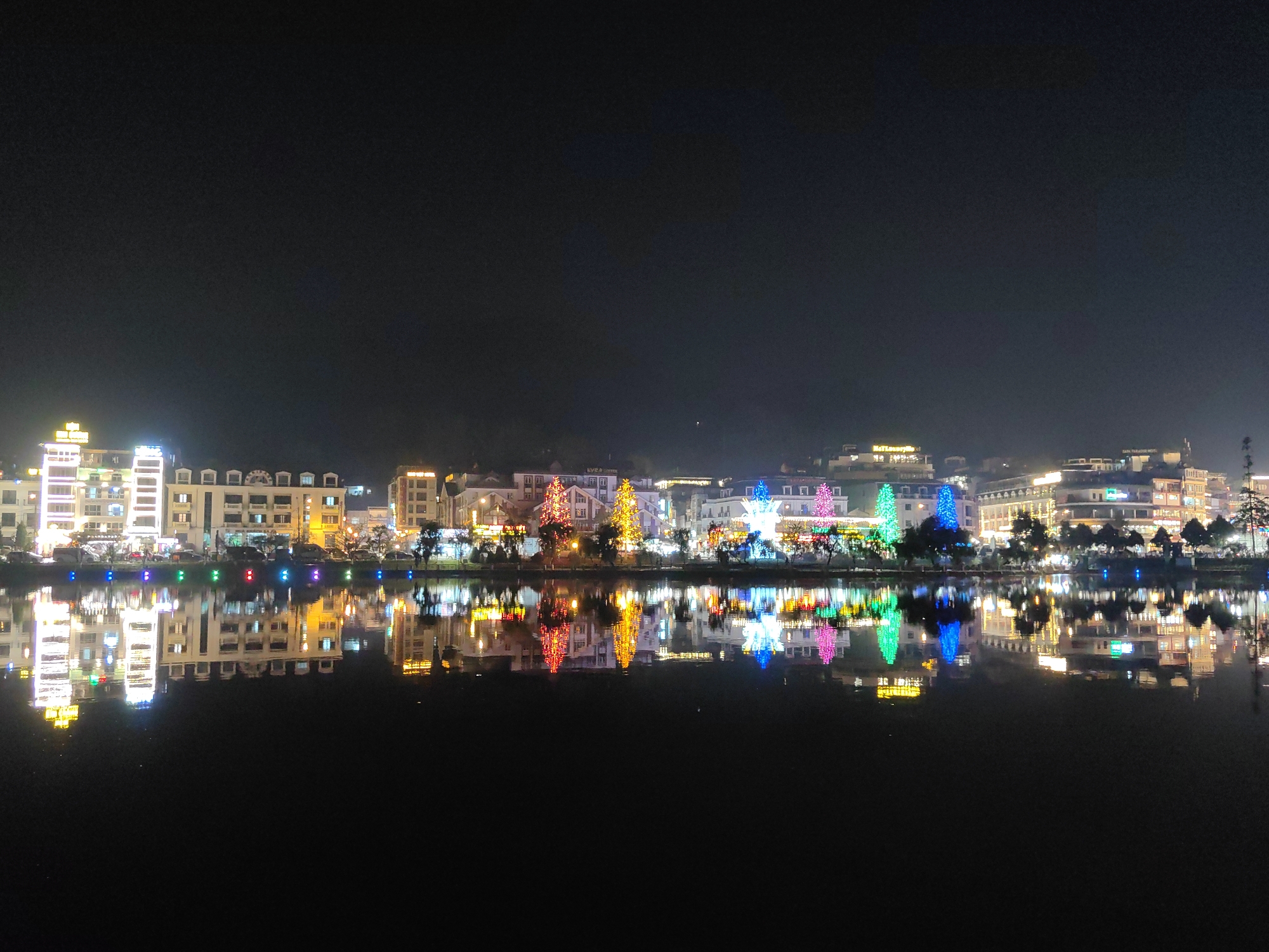 This image depicts a nighttime panoramic view of a section of Sa Pa, Vietnam, overlooking a body of water. The scene is dominated by a brightly illuminated cityscape reflected on the calm, dark surface of the water, which occupies the lower half of the frame.
Along the waterfront, a continuous row of multi-story buildings is visible, primarily constructed with light-colored facades and featuring numerous illuminated windows. Several of these structures display prominent signage, including "HA LOANG HOTEL," "SAPA PARADISE HOTEL," and "EYED HOTEL," indicating commercial or hospitality establishments. Interspersed among the buildings and lining the immediate edge of the water are numerous trees. Several of these trees are extensively decorated with multi-colored string lights, displaying hues of red, yellow, green, blue, and pink. A distinct row of circular, multi-colored lights (blue, green, red, yellow) is positioned directly at the water's edge, enhancing the festive illumination. The upper portion of the image shows a dark, cloudless night sky. No individuals or specific actions are discernible within the frame.