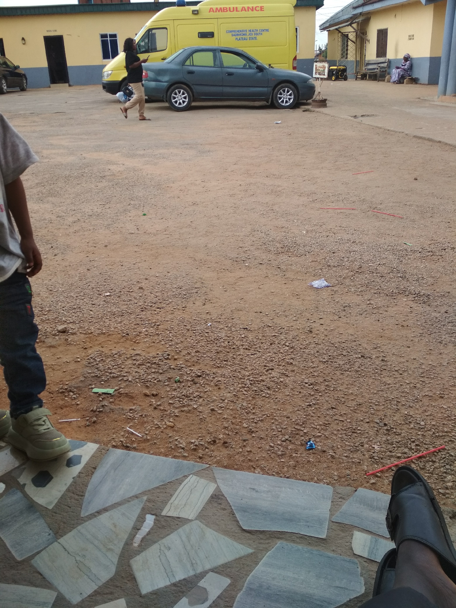 The image depicts an outdoor scene. A yellow ambulance and a dark-colored sedan are parked in a gravel parking lot. A person in dark clothing and tan pants is walking past the vehicles. An elderly woman is sitting on a bench in front of a building. In the foreground, a person is standing on a stone-tiled surface wearing green sneakers and blue jeans, while another person sits with black sandals. The location is the Comprehensive Health Centre Dadinkowa Jos South Plateau State.