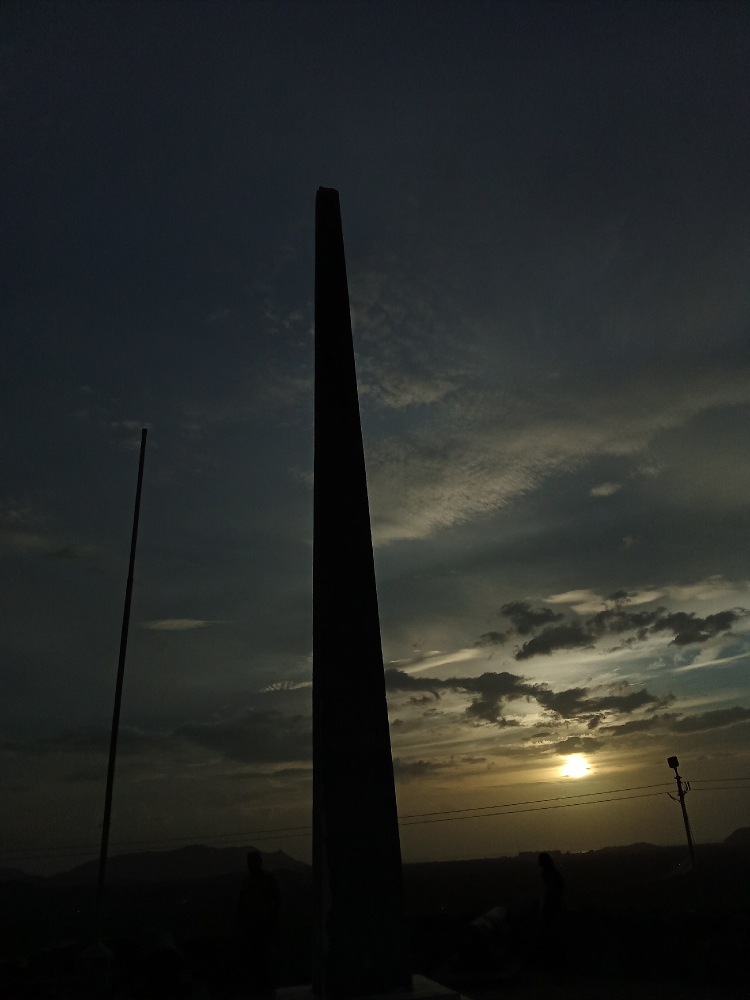 The photograph depicts a tall, dark obelisk silhouetted against a sky with a setting sun visible through the clouds. A thinner pole-like structure stands to the left of the obelisk. In the foreground, dark figures are visible, possibly people observing the scene. In the background, a landscape of dark hills and a utility pole with wires can be discerned against the horizon. The location is purported to be in the city of Santa Ana, Venezuela.