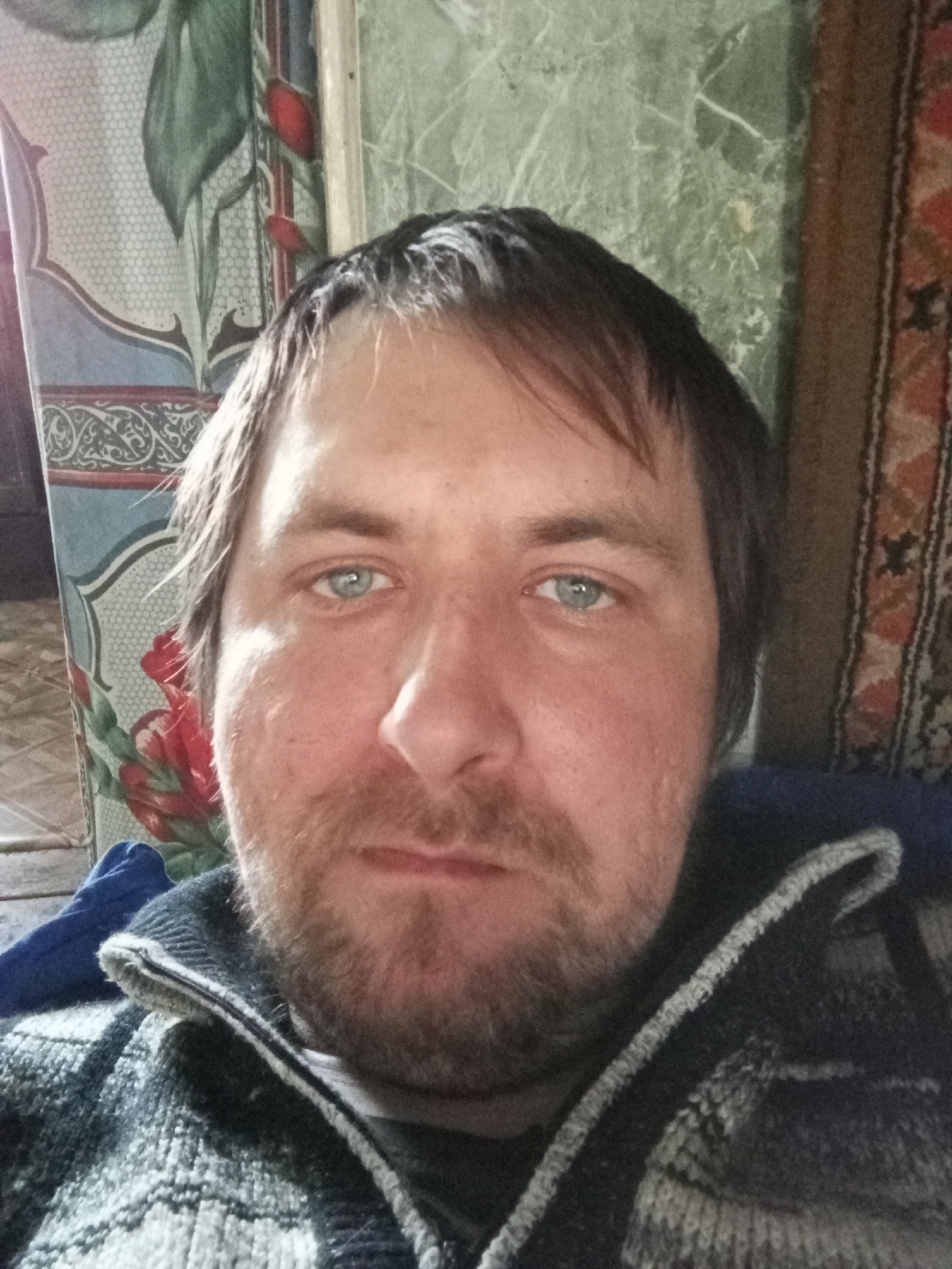 A selfie of a middle-aged man with fair skin, light blue eyes, and brown hair. He has a mustache and a short beard. He is wearing a zipped-up, dark-colored sweater with a light-colored pattern and trim. Behind him are two different patterned fabric panels, one featuring a floral design with red roses, and the other a more geometric, traditional pattern. The location is indicated as Parabel, Russia.